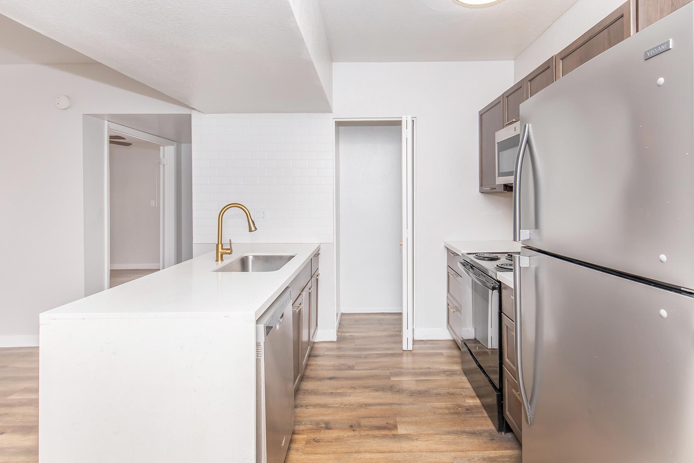 Modern kitchen featuring stainless steel appliances, including a refrigerator, stove, and dishwasher. Light-colored cabinetry and a white countertop complement the space, which has a warm wooden floor. The open layout connects to a dining area with ample natural light.