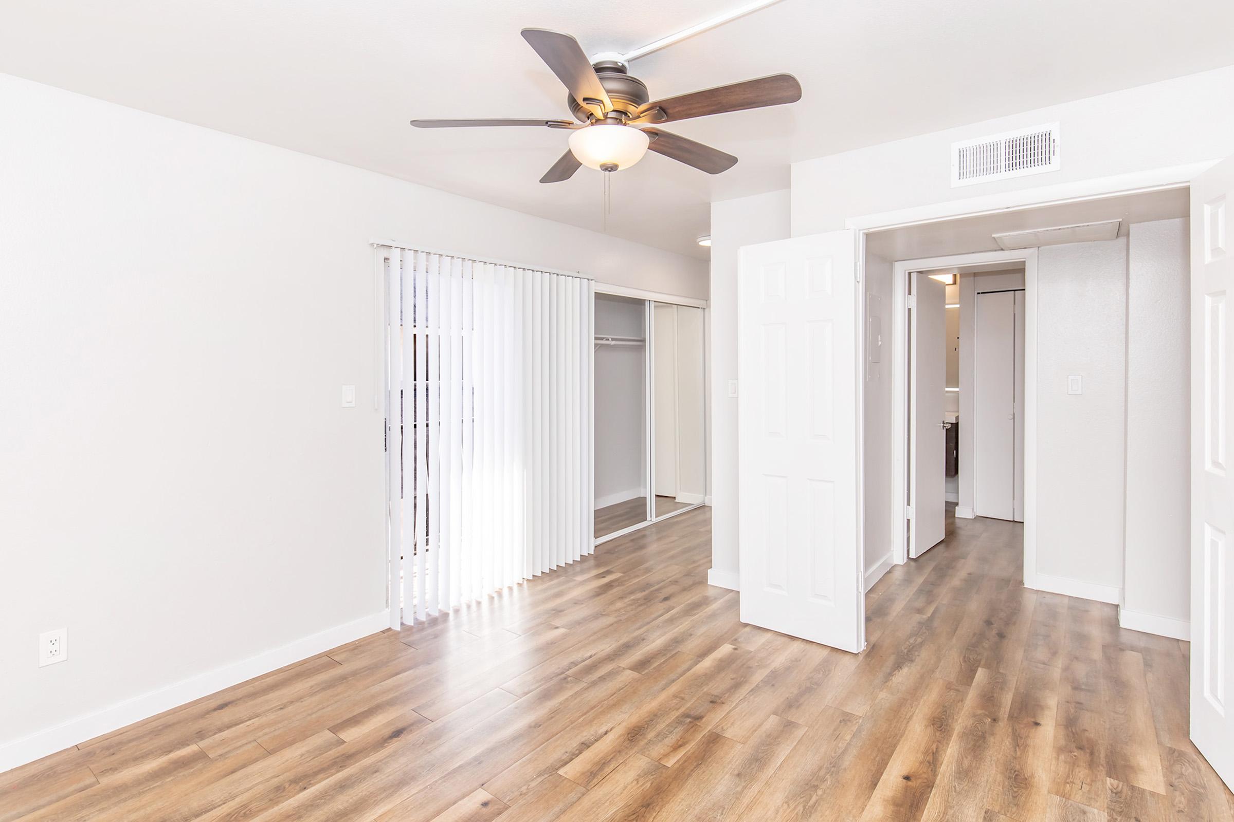 Spacious room with light-colored walls and wood laminate flooring. A ceiling fan is visible, and there are white vertical blinds covering a window. To the left, there is a closet with sliding doors, and an open doorway leads to a hallway. The overall atmosphere is bright and airy.