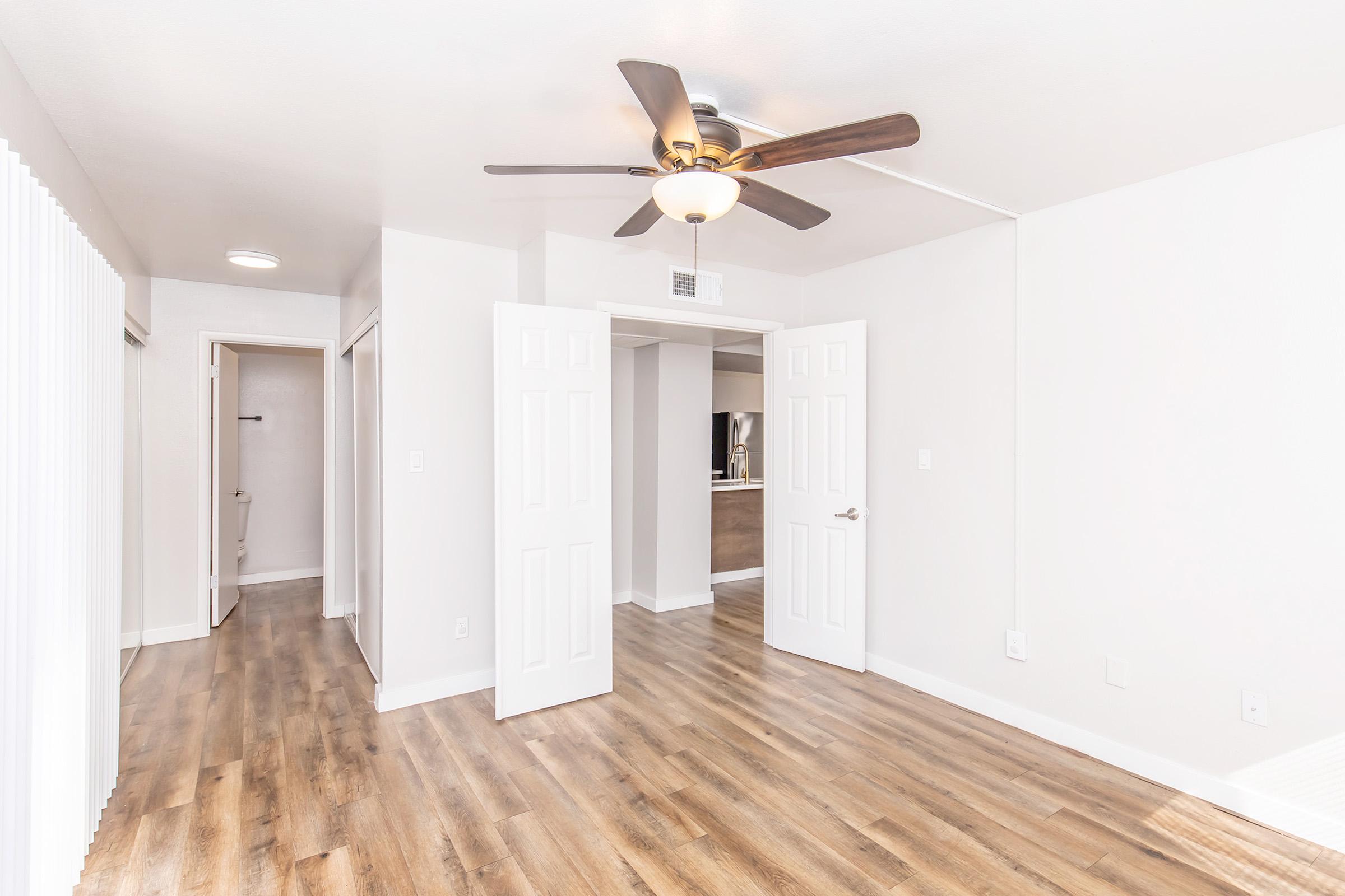 A spacious, well-lit room featuring light-colored walls and wooden flooring. Two open doors lead to adjacent areas, and a ceiling fan is visible. The layout conveys a modern and inviting atmosphere, suitable for various uses like a living area or bedroom.