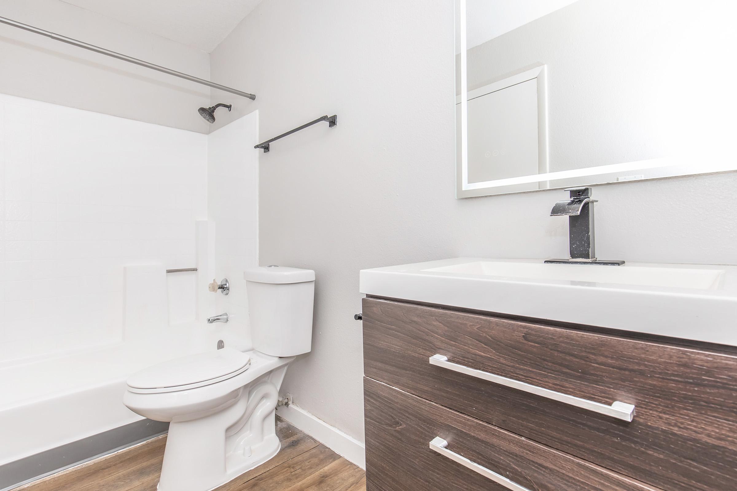 A modern bathroom featuring a white bathtub with a shower, a toilet, and a sleek dark wooden vanity with a white countertop and a mirror above. The walls are painted in a light color, and the floor is styled with laminate wood. The space is illuminated with natural light.