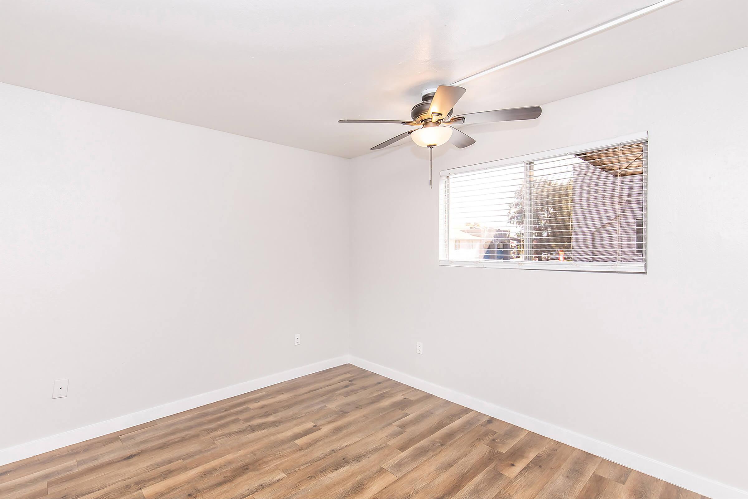 Empty room with light-colored walls and wooden flooring. A ceiling fan is mounted in the center, and a window with blinds allows natural light to enter. The overall atmosphere is bright and open, suitable for various uses.