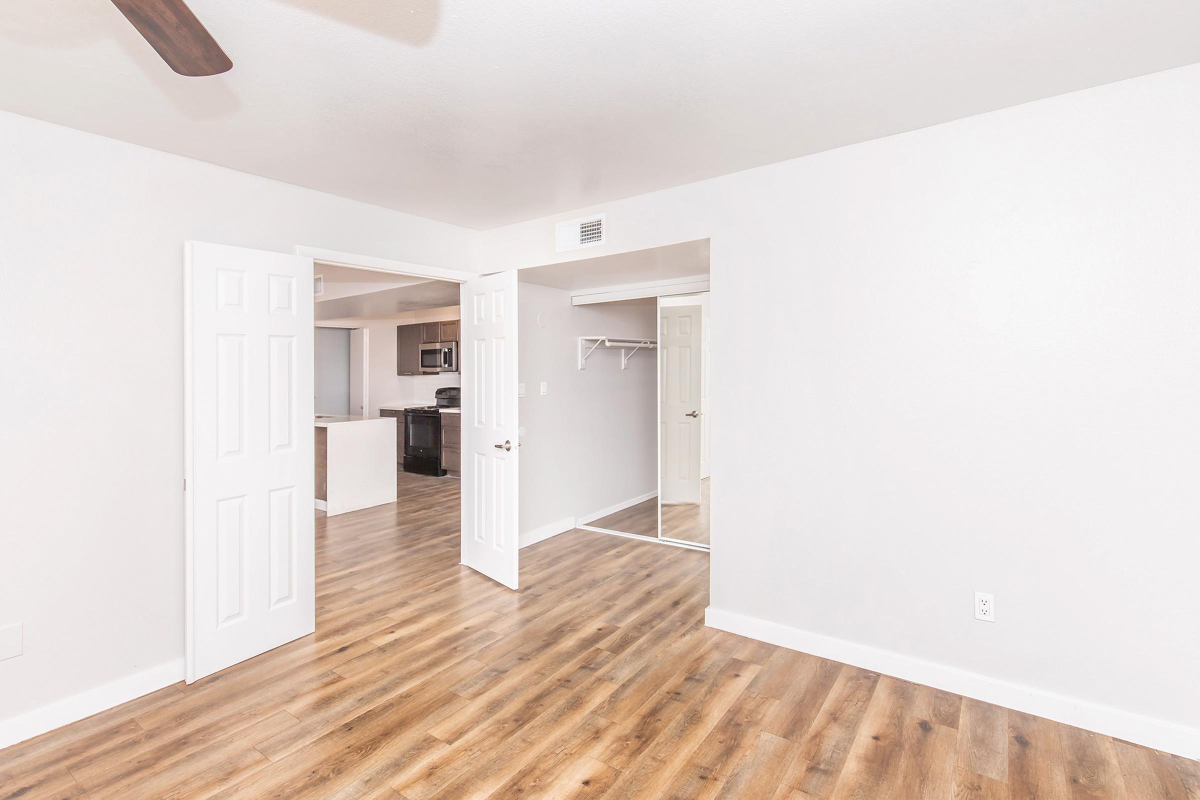 Interior view of a room with light-colored walls and wooden flooring. Two open doors lead to a potential closet area and a kitchen/living space in the background. The room appears bright and spacious, with a ceiling fan visible. Simple, modern design aesthetic.