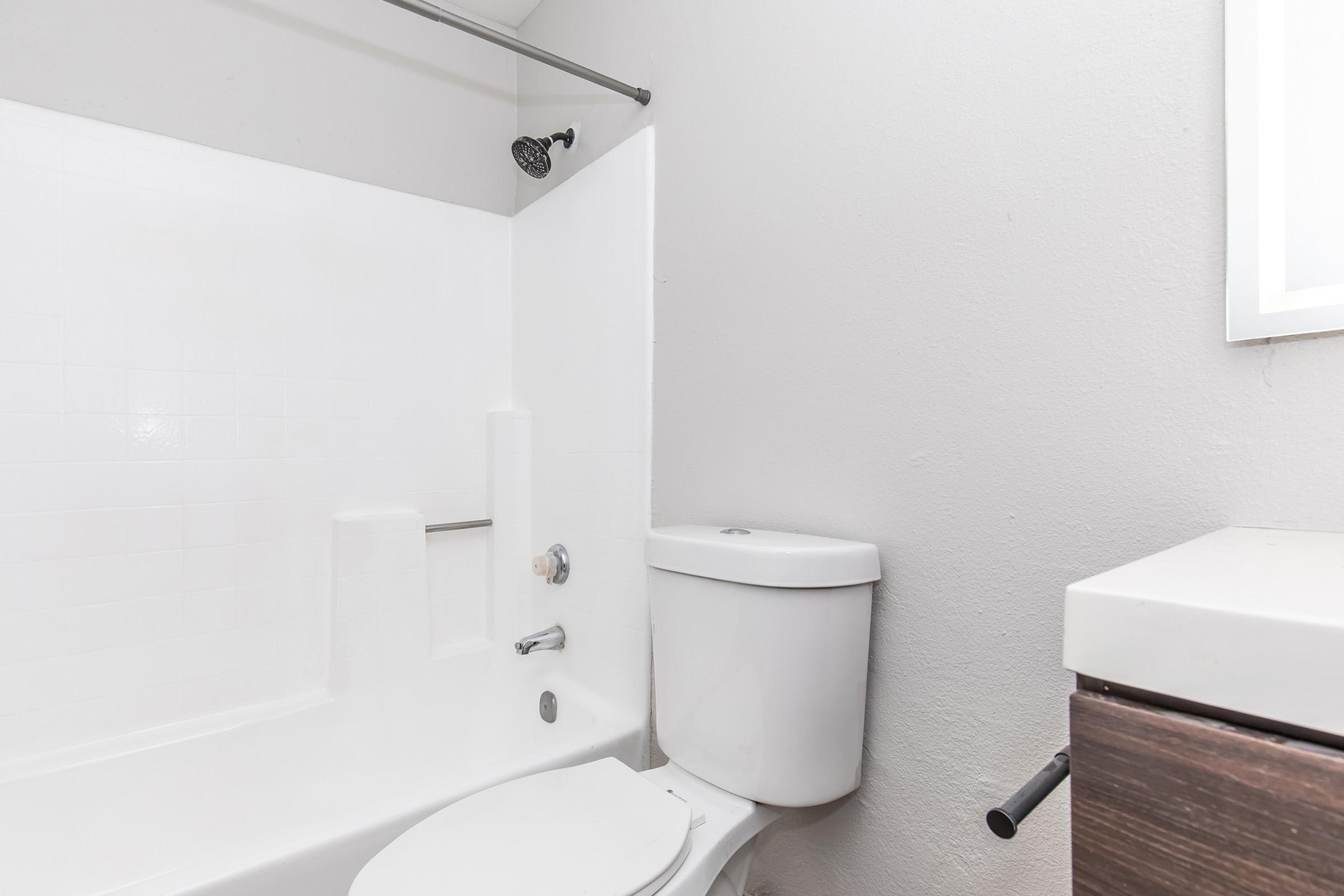 A clean, minimalist bathroom featuring a white bathtub with a shower head, a white toilet, and a small vanity with a dark wood finish. The walls are painted light gray, and a window allows natural light to enter the space.
