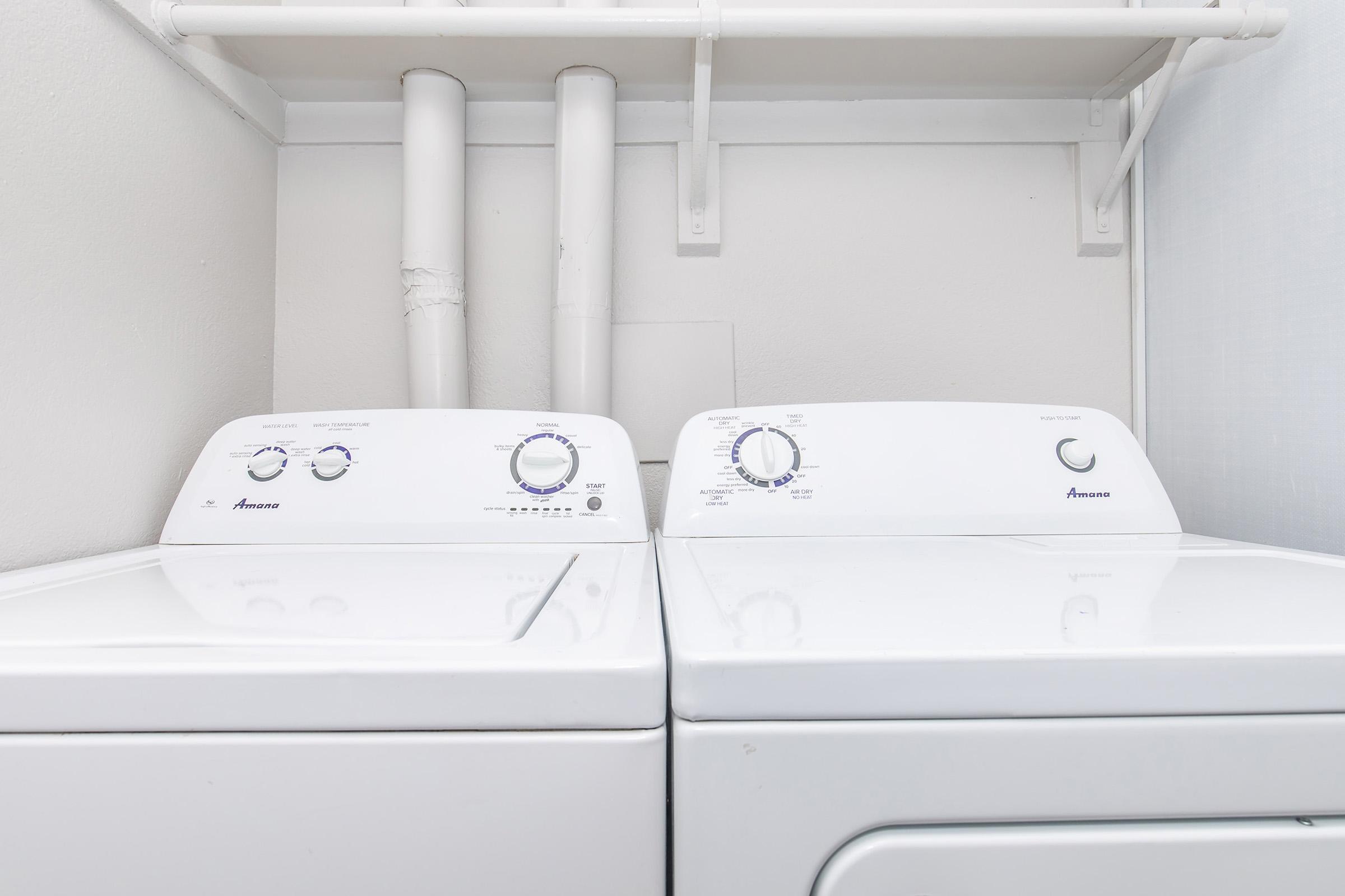 A white washing machine and dryer side by side, located in a small laundry area. The machines have control dials and knobs on the front. The space is clean and the walls are painted a light color, with white pipes running above the appliances.