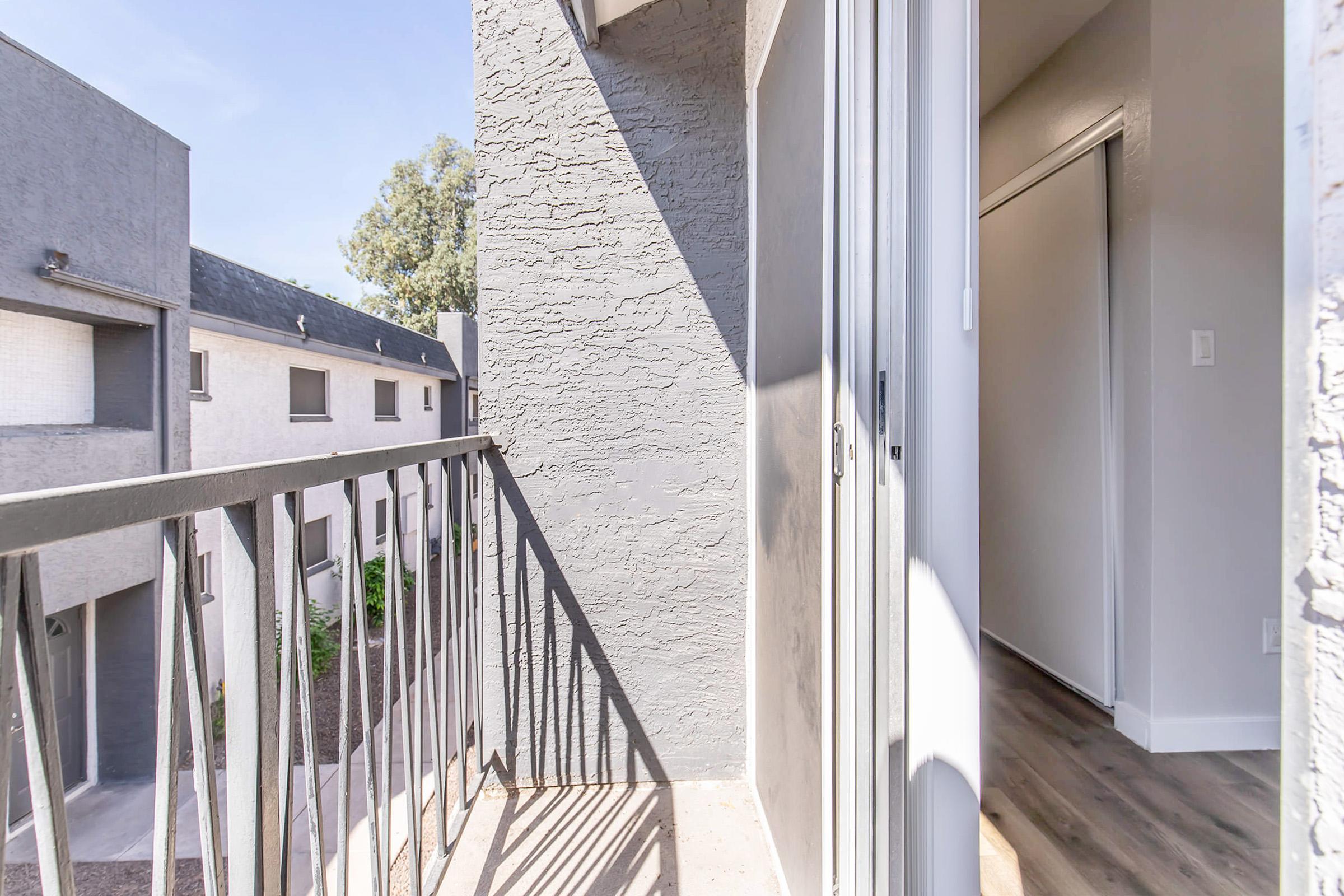View from a balcony showing a small outdoor space with a metal railing. The scene includes light-colored walls of adjacent buildings and a glimpse of greenery below. Sunlight creates a bright atmosphere, highlighting the textures of the balcony and surrounding areas.