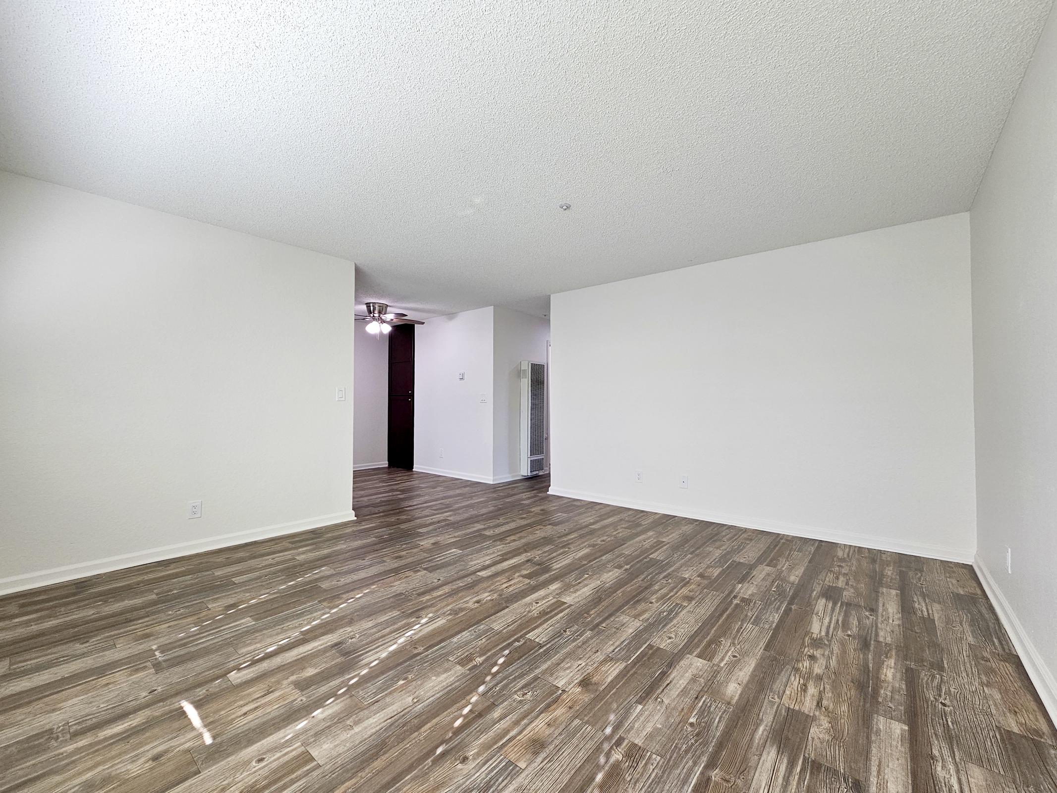 A spacious, empty room with a light-colored textured ceiling, vinyl wood flooring, and white walls. There is a ceiling fan visible and an open doorway leading to another area. The room is well-lit with natural light, creating an inviting atmosphere.