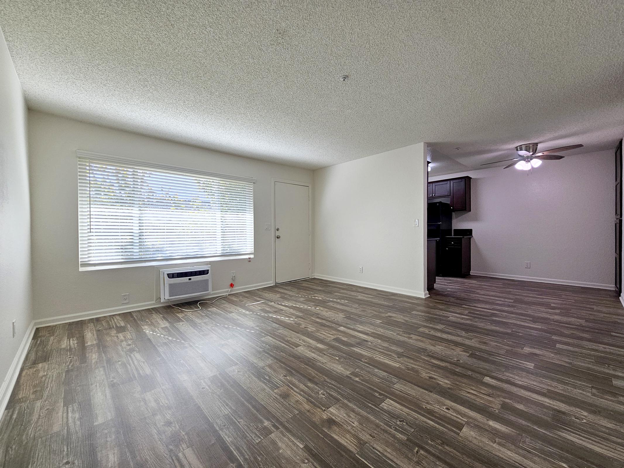A spacious living area with wood-style flooring, a large window allowing natural light, and an air conditioning unit. To the right, there is a doorway leading to a kitchen area with dark cabinetry. The walls are painted in a light color, creating a bright and open atmosphere.