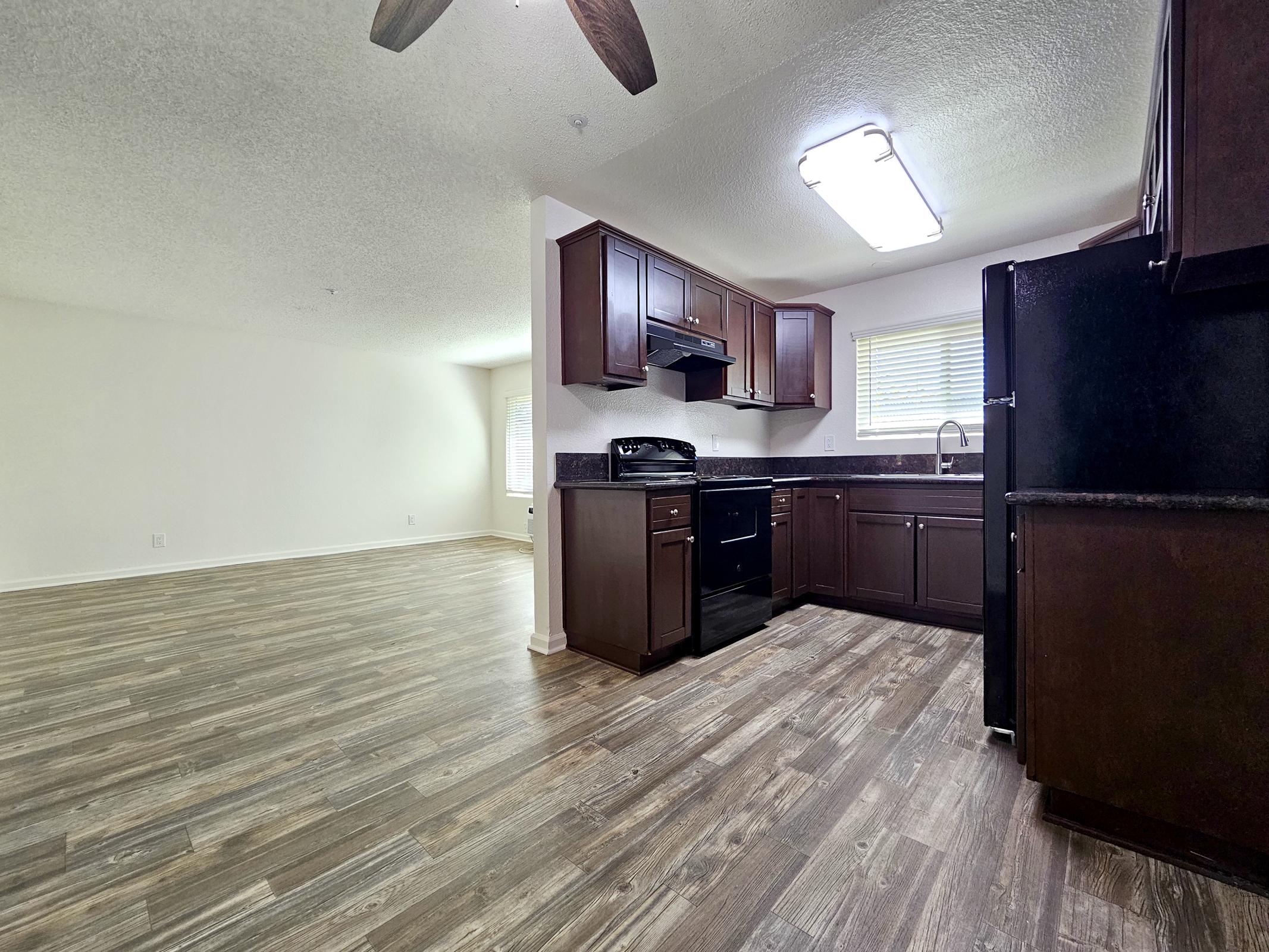 A modern kitchen featuring dark wood cabinetry, a black refrigerator, and a sink with a window above it. The open layout includes a spacious living area with light-colored walls and a ceiling fan. The flooring is a wood-like laminate, providing a warm and inviting atmosphere.