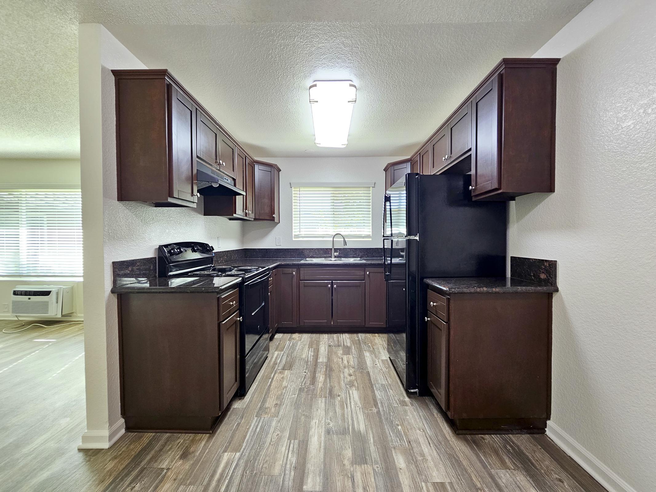 A modern kitchen featuring dark wooden cabinets, a black stove, and a black refrigerator. The countertops are dark, and there's a sink beneath a window that lets in natural light. The flooring is a light wood grain, contributing to a warm atmosphere.
