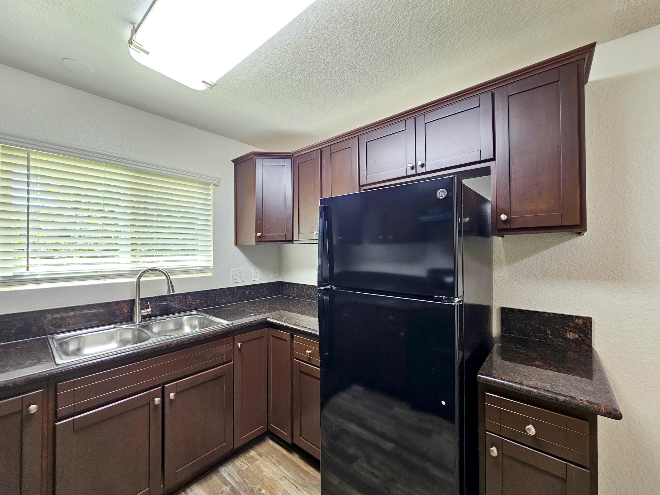 A modern kitchen with dark wood cabinets, a black refrigerator, and a double sink. The countertop is dark, and there is a window with blinds allowing natural light to enter. The overall design is sleek and contemporary, featuring a well-organized layout.