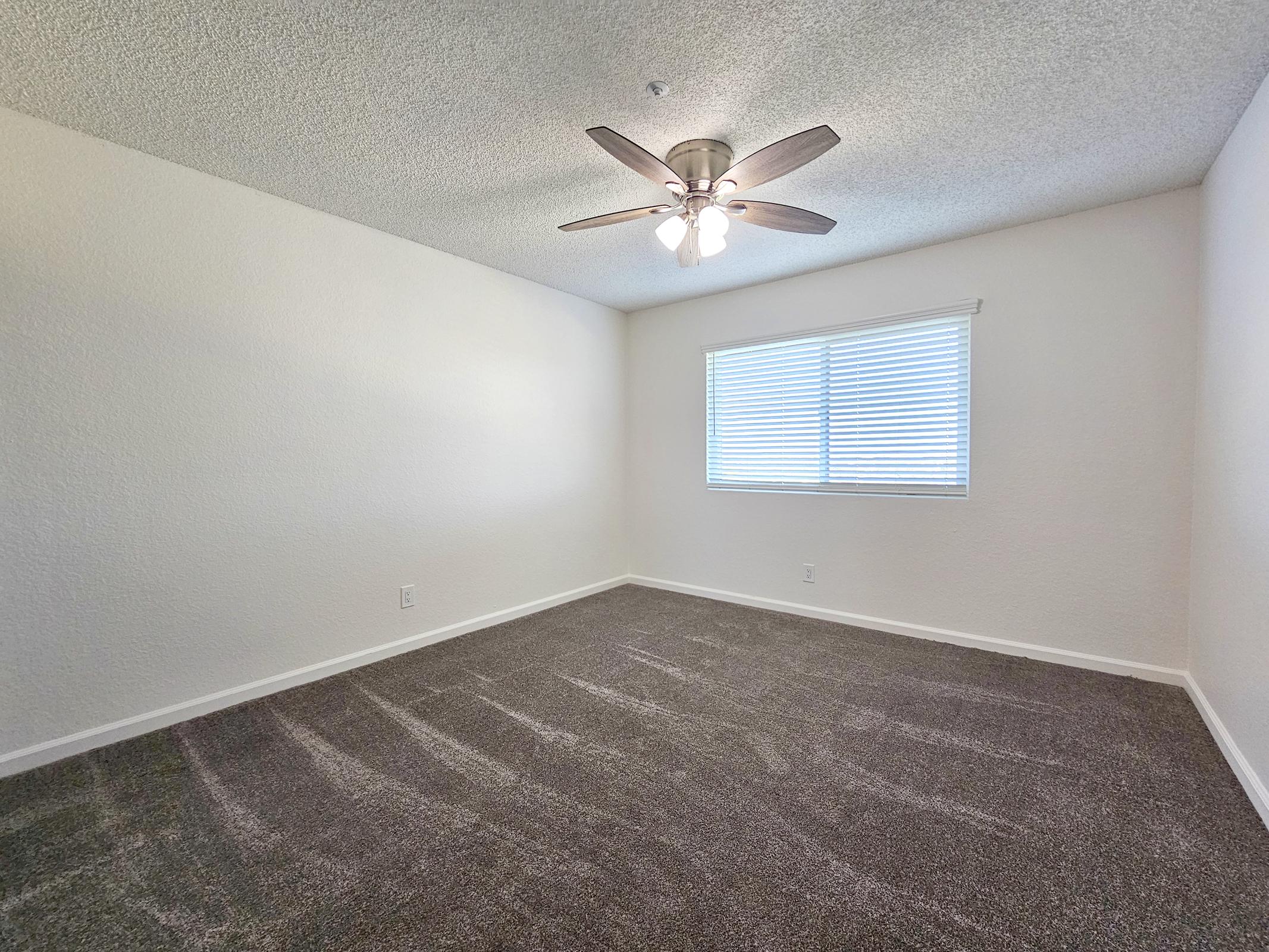 A spacious, empty room with light brown carpet and a ceiling fan. There is a window with blinds allowing natural light to enter, and the walls are painted white. The room appears clean and well-maintained, suitable for use as a bedroom or office.