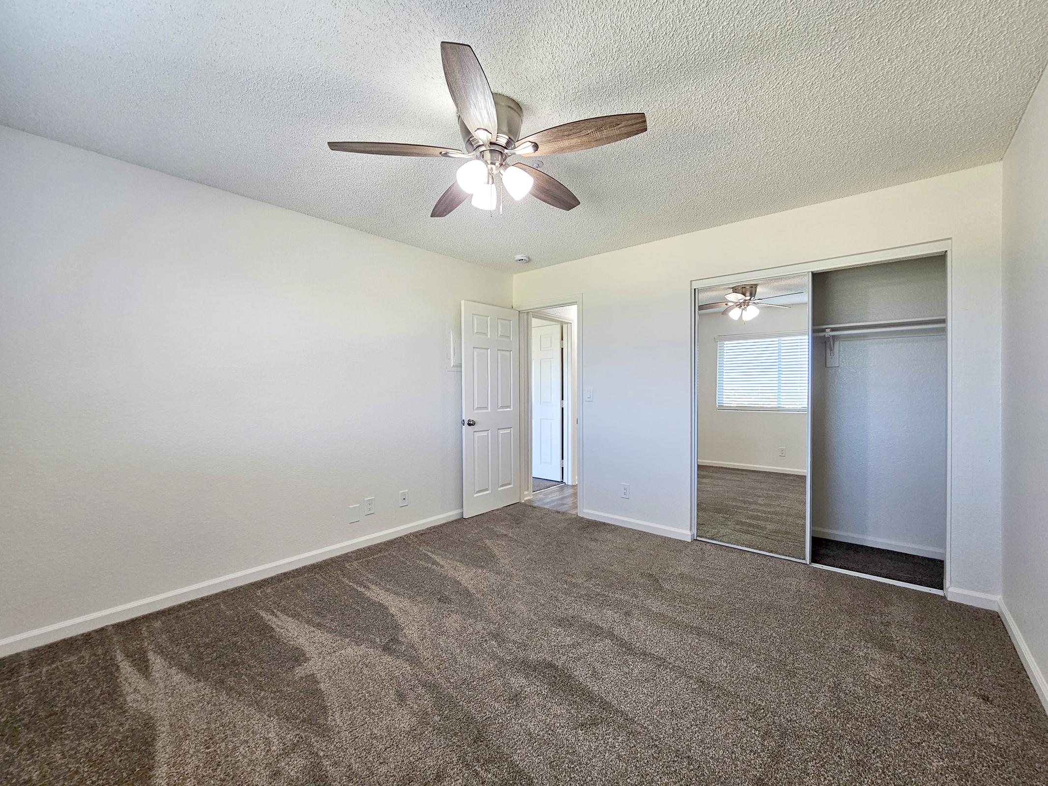 A spacious, empty room featuring light-colored walls and a ceiling fan with wood blades. The floor is covered with brown carpet. There's a mirrored sliding closet door on one side and a doorway leading to another room. Natural light enters through a window, creating a bright atmosphere.