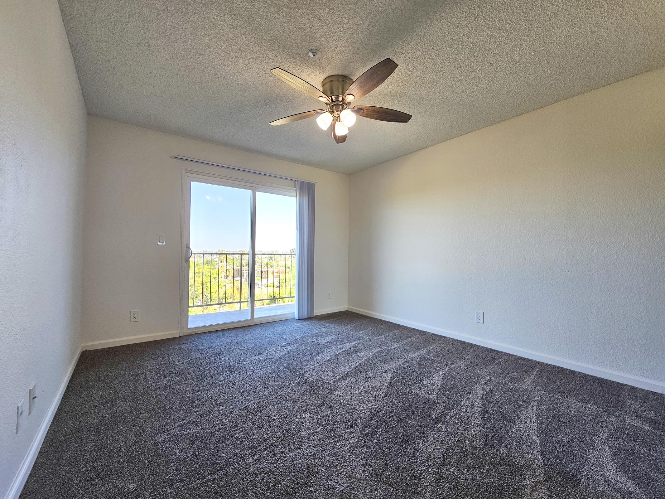 A bright, empty room featuring light beige walls and plush gray carpet. A ceiling fan with wooden blades hangs overhead. Large sliding glass doors lead to a balcony with a view, allowing natural light to fill the space. The overall atmosphere is open and inviting, suitable for various furniture arrangements.
