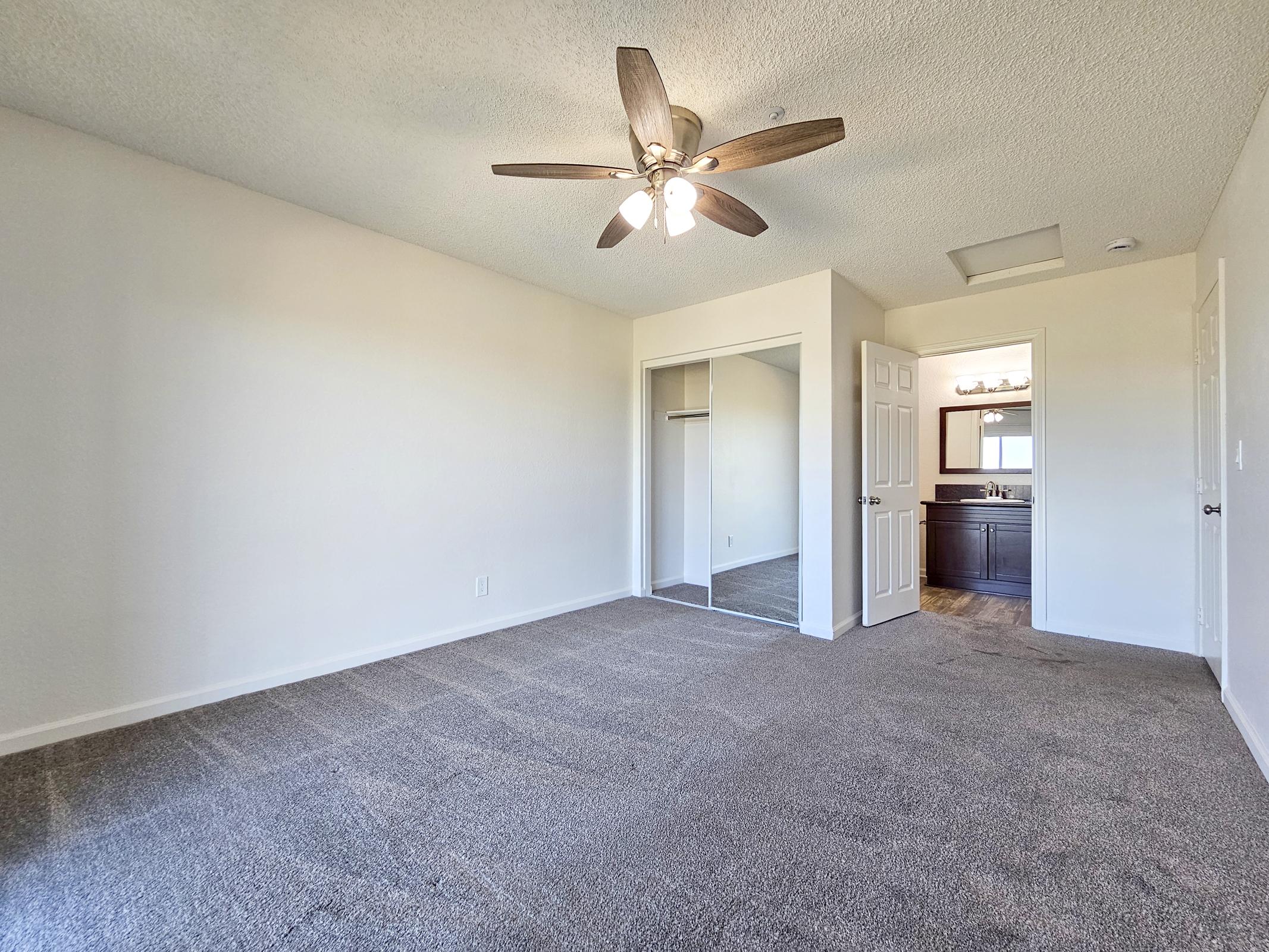 A bright, empty bedroom featuring light-colored walls, a ceiling fan with a light fixture, and a large carpeted area. To one side, there's a mirrored closet, and in the back, a doorway leads to a separate bathroom area with dark cabinetry. The room has natural light coming in from a window.