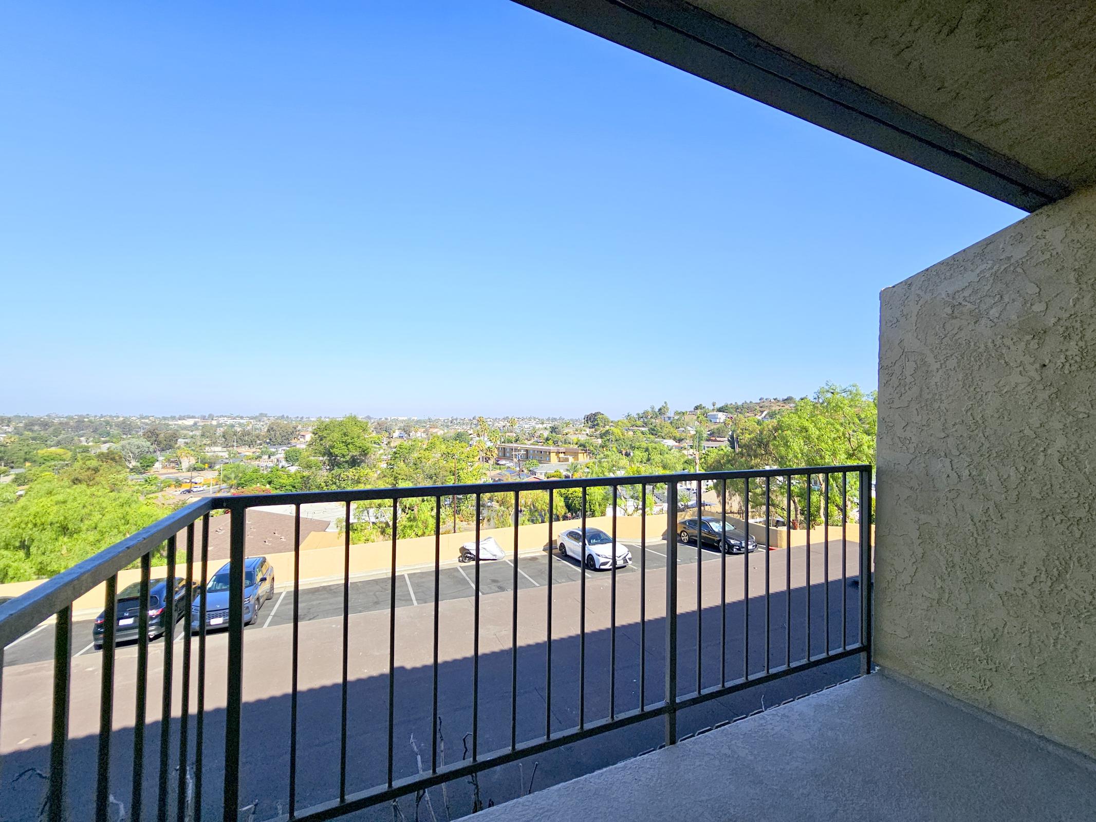 A view from a balcony overlooking a residential area with cars parked below. The clear blue sky stretches above, and green trees are visible in the distance, indicating a pleasant, sunny day. The scene captures a tranquil suburban landscape.
