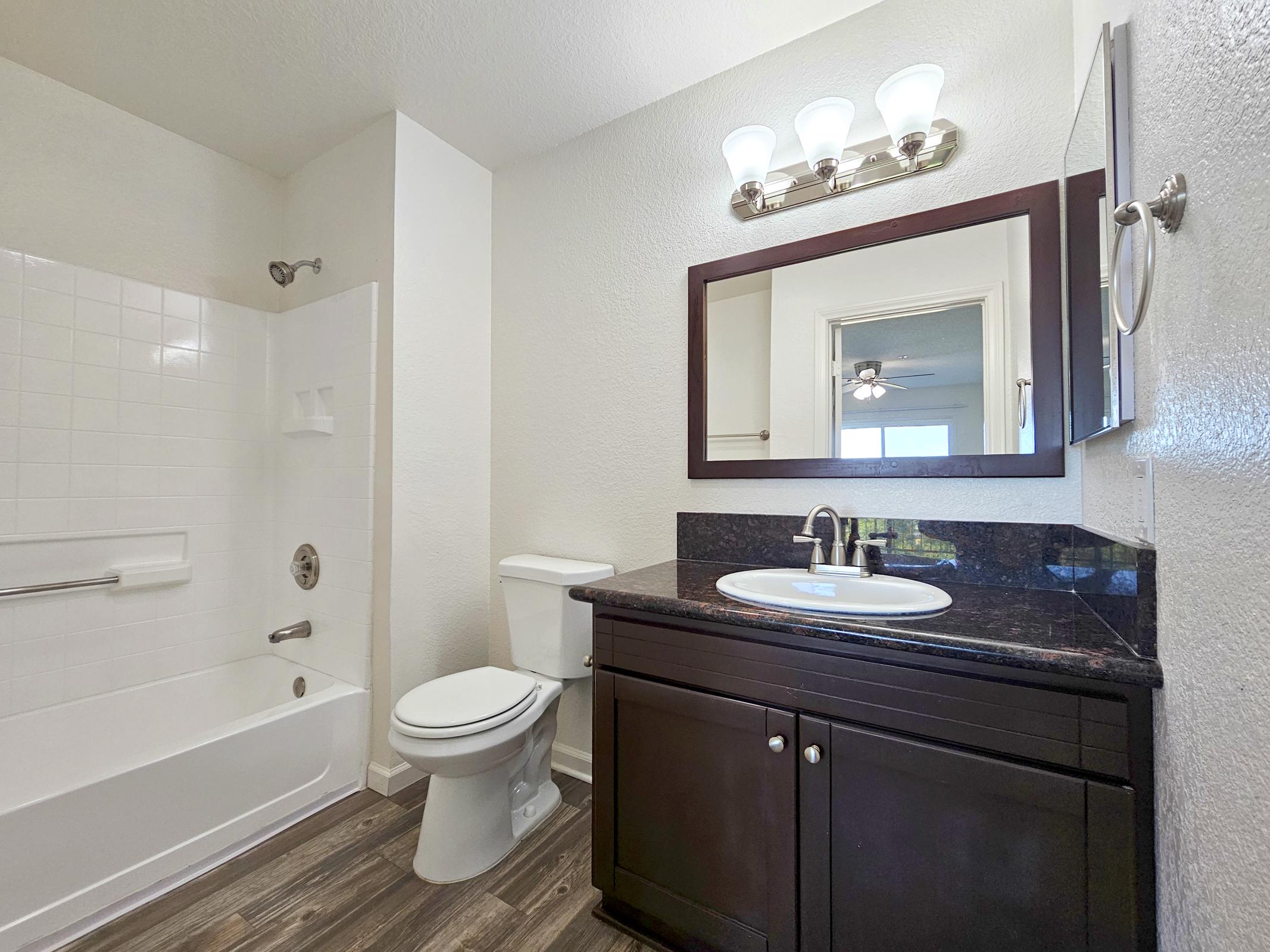 A modern bathroom featuring a white bathtub with a showerhead, a dark wood vanity with a round sink and granite countertop, a toilet, and a wall-mounted mirror. Ceiling light fixtures illuminate the space, while vinyl flooring adds a contemporary touch. A window reflects natural light.
