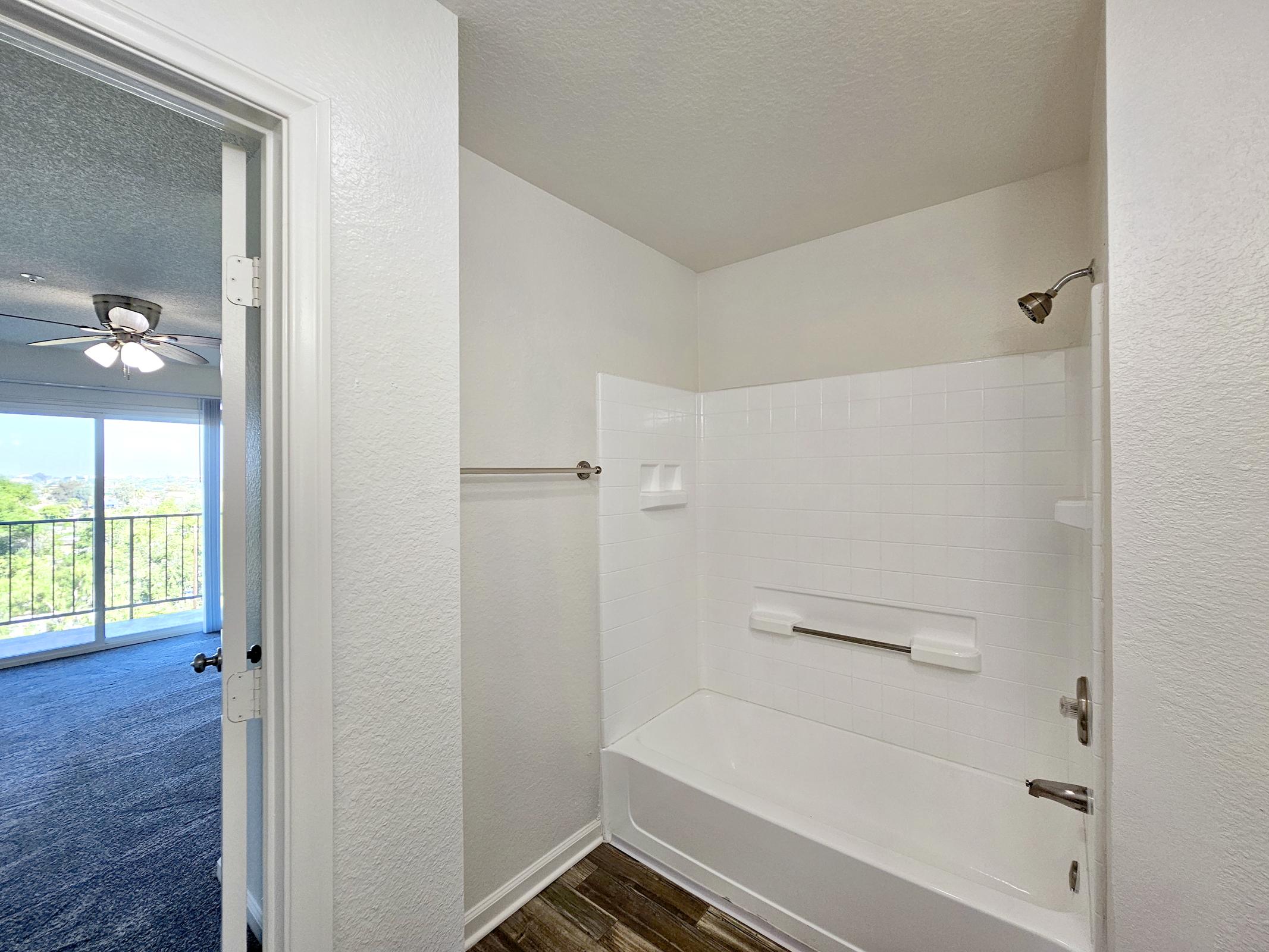 Clean bathroom with a white tub and shower, featuring a grab bar and wall-mounted soap dish. A door leads to a bright, spacious area with a large window overlooking a scenic view. The flooring is a wood-like finish, and there's a ceiling fan visible in the background.