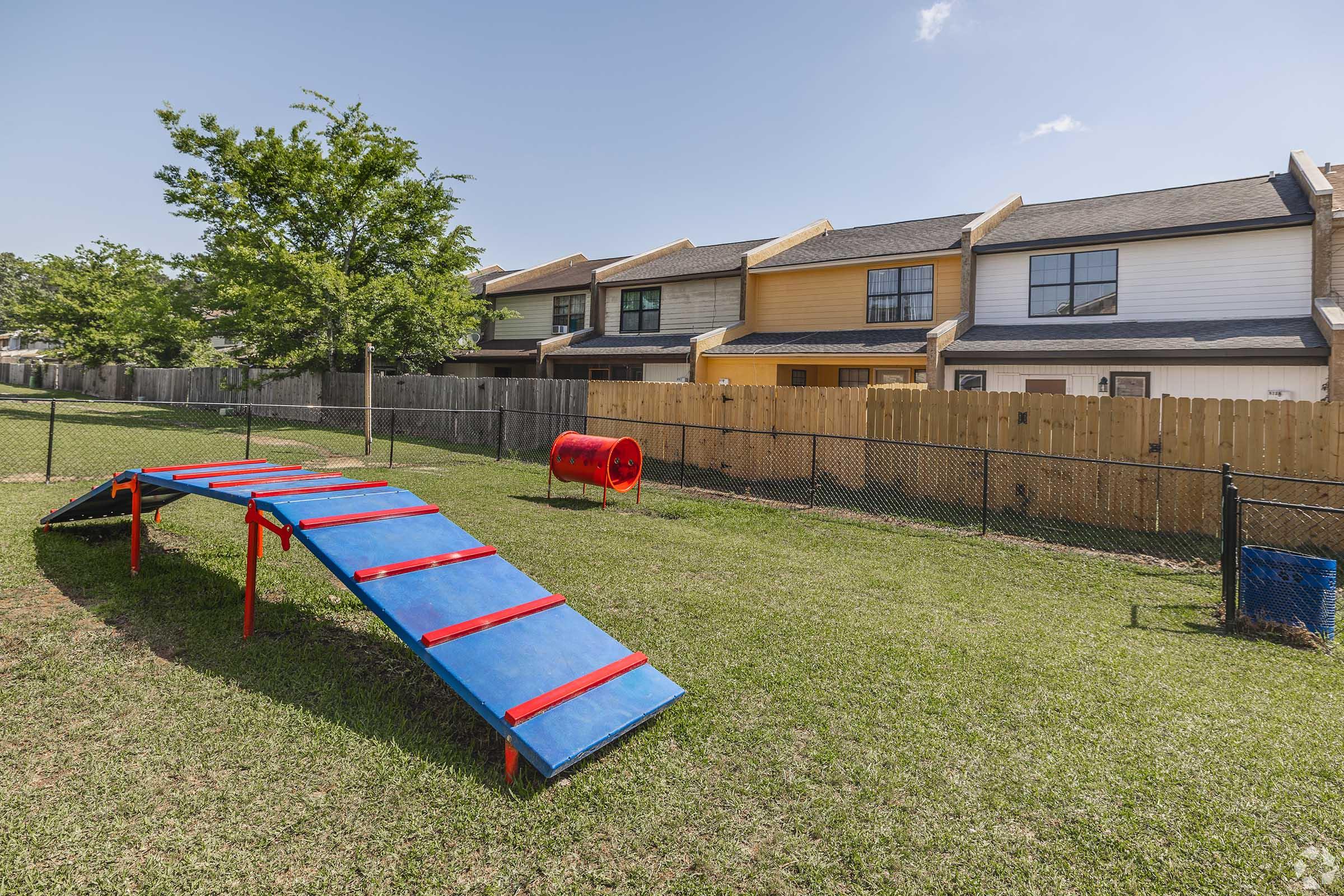 A bright and sunny backyard featuring a small agility training setup for dogs, consisting of a blue ramp with red accents and a red tunnel. In the background, there are several residential houses with a wooden fence and green grass. The sky is clear with a few clouds.