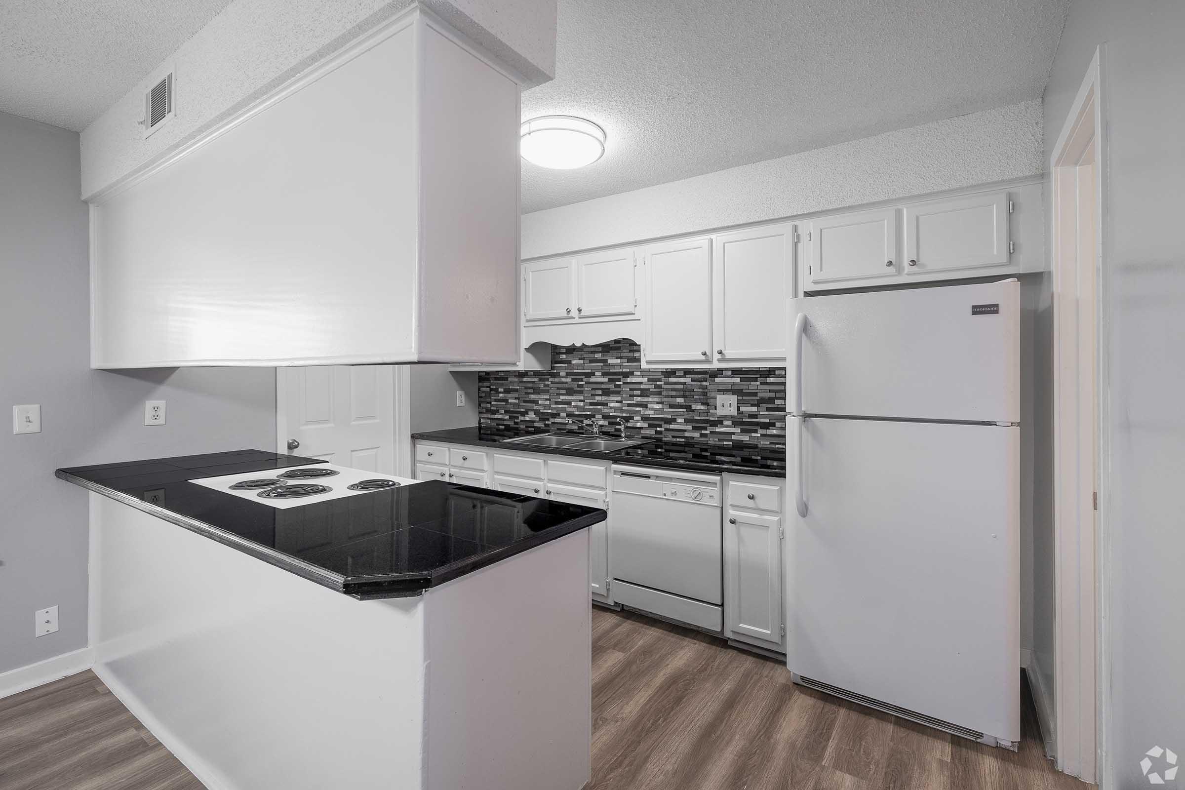 Modern kitchen featuring a black countertop, white cabinetry, an electric stove, and a stainless steel dishwasher. The backsplash is made up of grey and black tiles. A white refrigerator stands against the wall, and the space is well-lit with a ceiling light. The flooring is wood-like laminate.