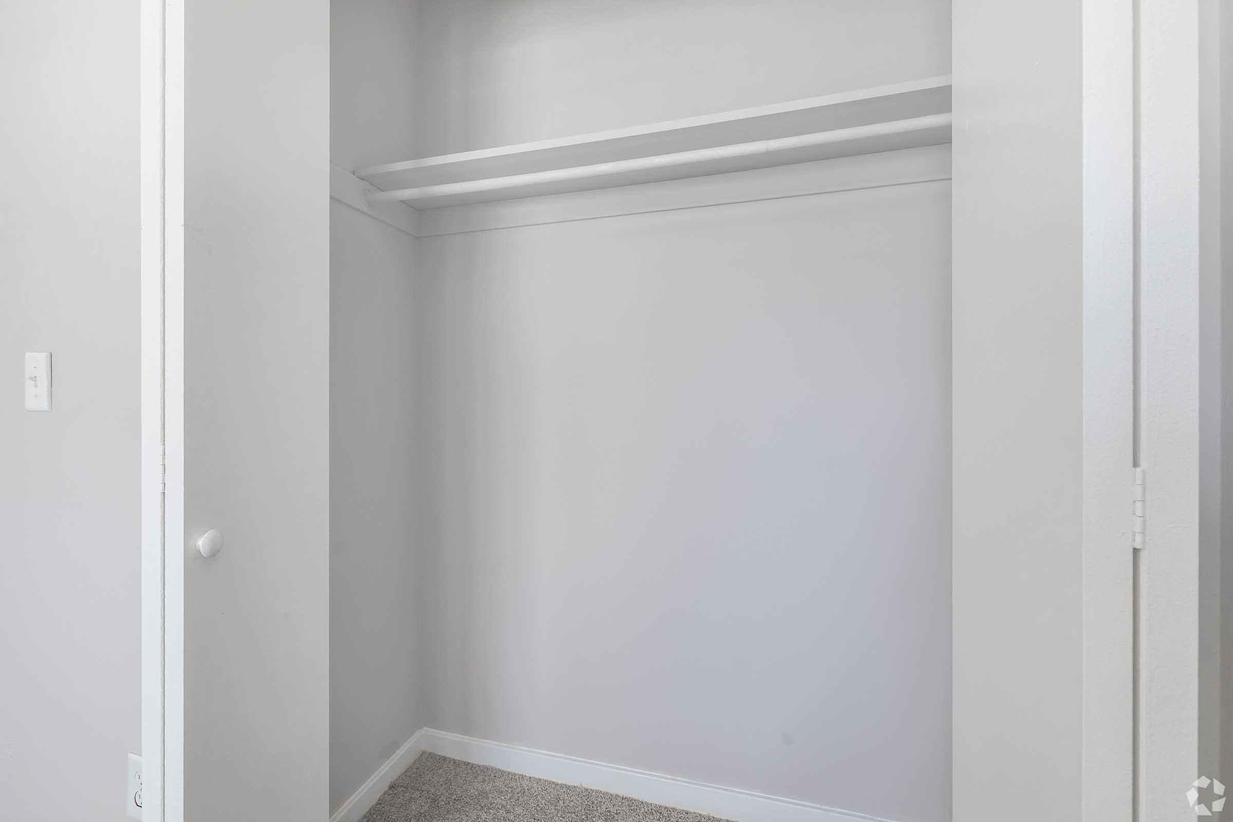 Empty closet with white walls and carpeted floor. Two shelves are visible at the top, and there is a door on the right side. The space is well-lit, creating a clean and minimalistic appearance.
