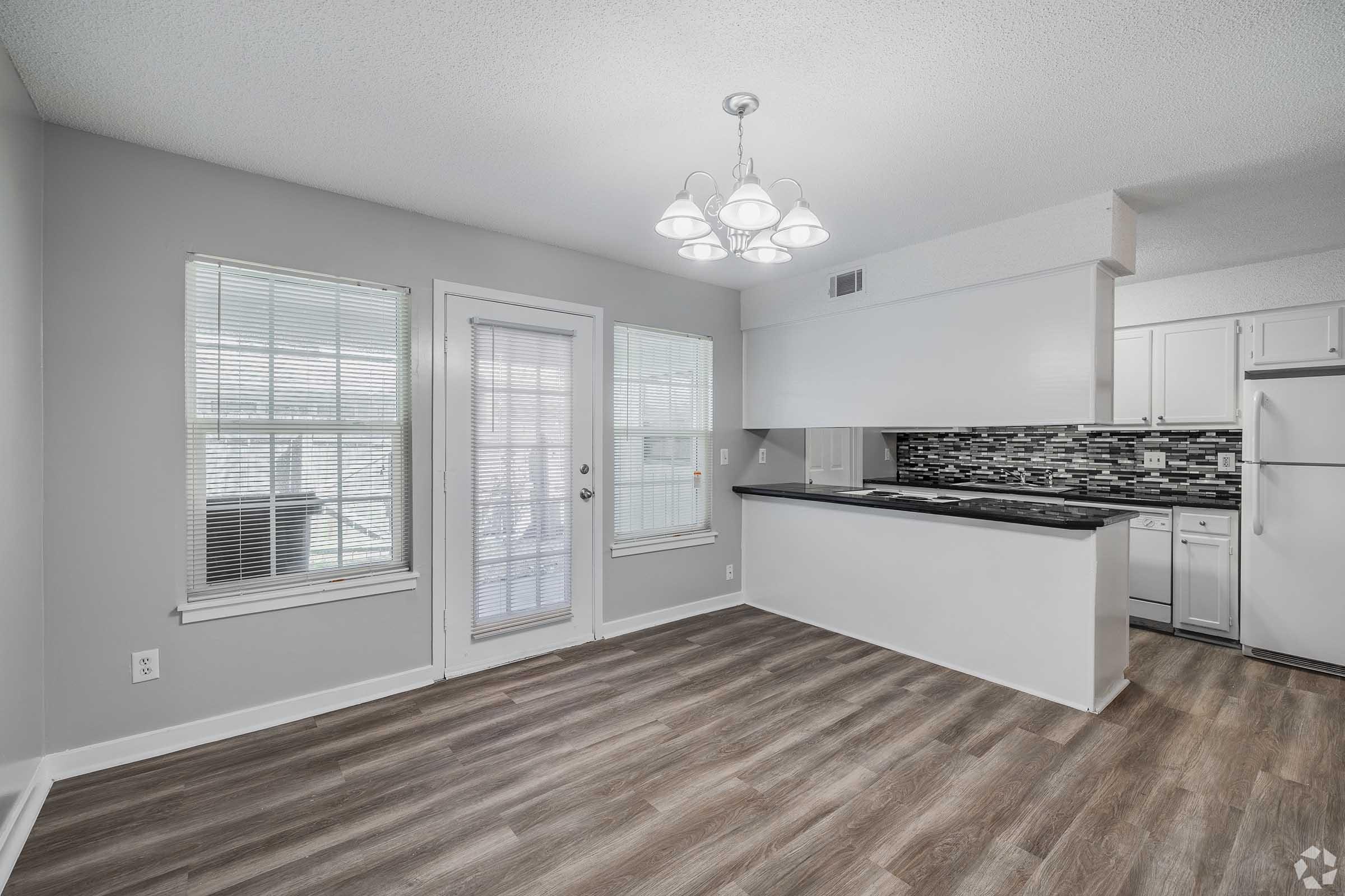 A modern kitchen and dining area featuring a pendant light, large windows with natural light, and a sliding glass door. The kitchen includes sleek cabinets and a backsplash with a mosaic design. The flooring is wood-like, adding warmth to the space.