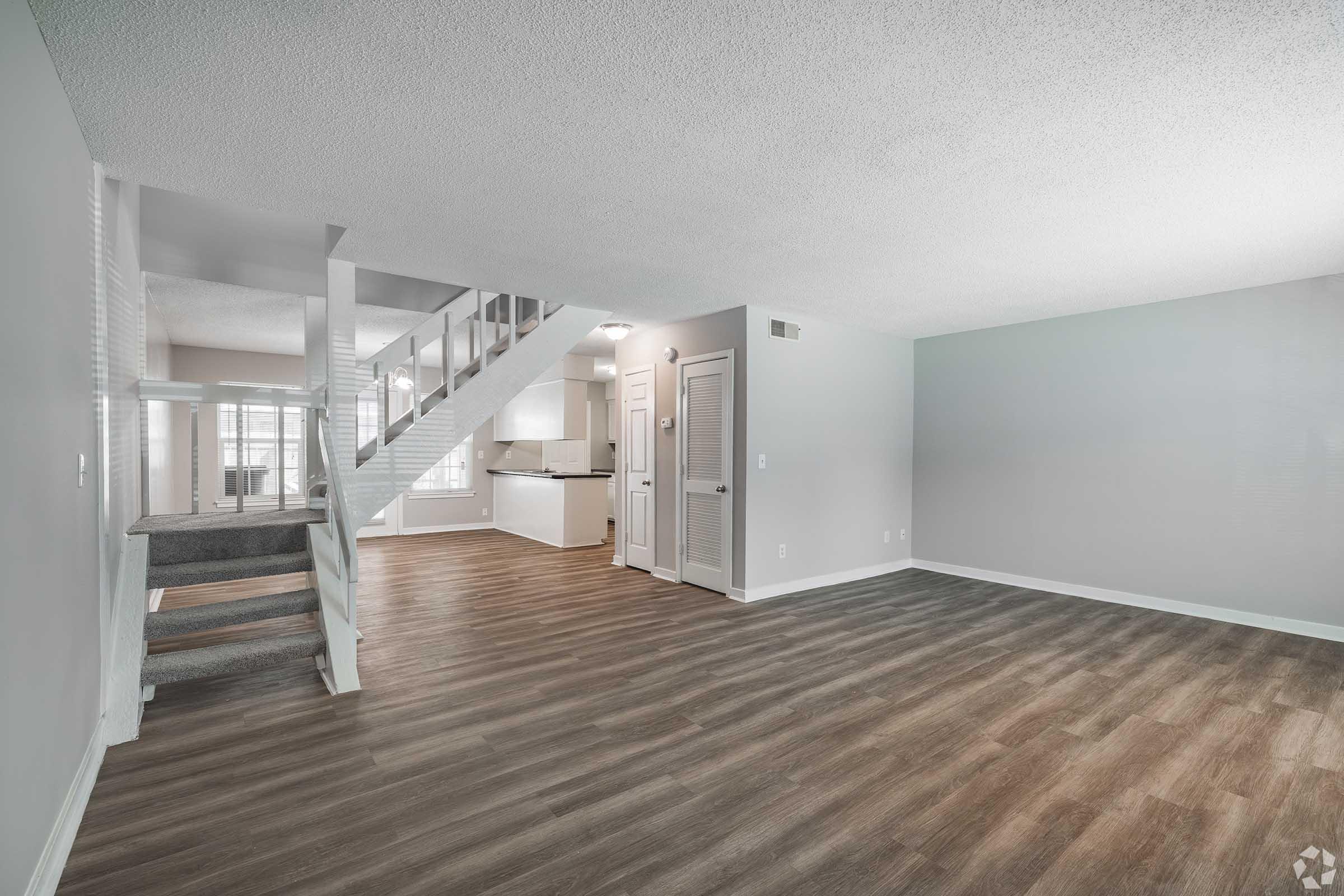 Spacious living area featuring a light gray color scheme, hardwood-style flooring, and a staircase leading to an upper level. The open floor plan connects to a modern kitchen visible in the background. Natural light filters through large windows, creating a bright and inviting atmosphere.