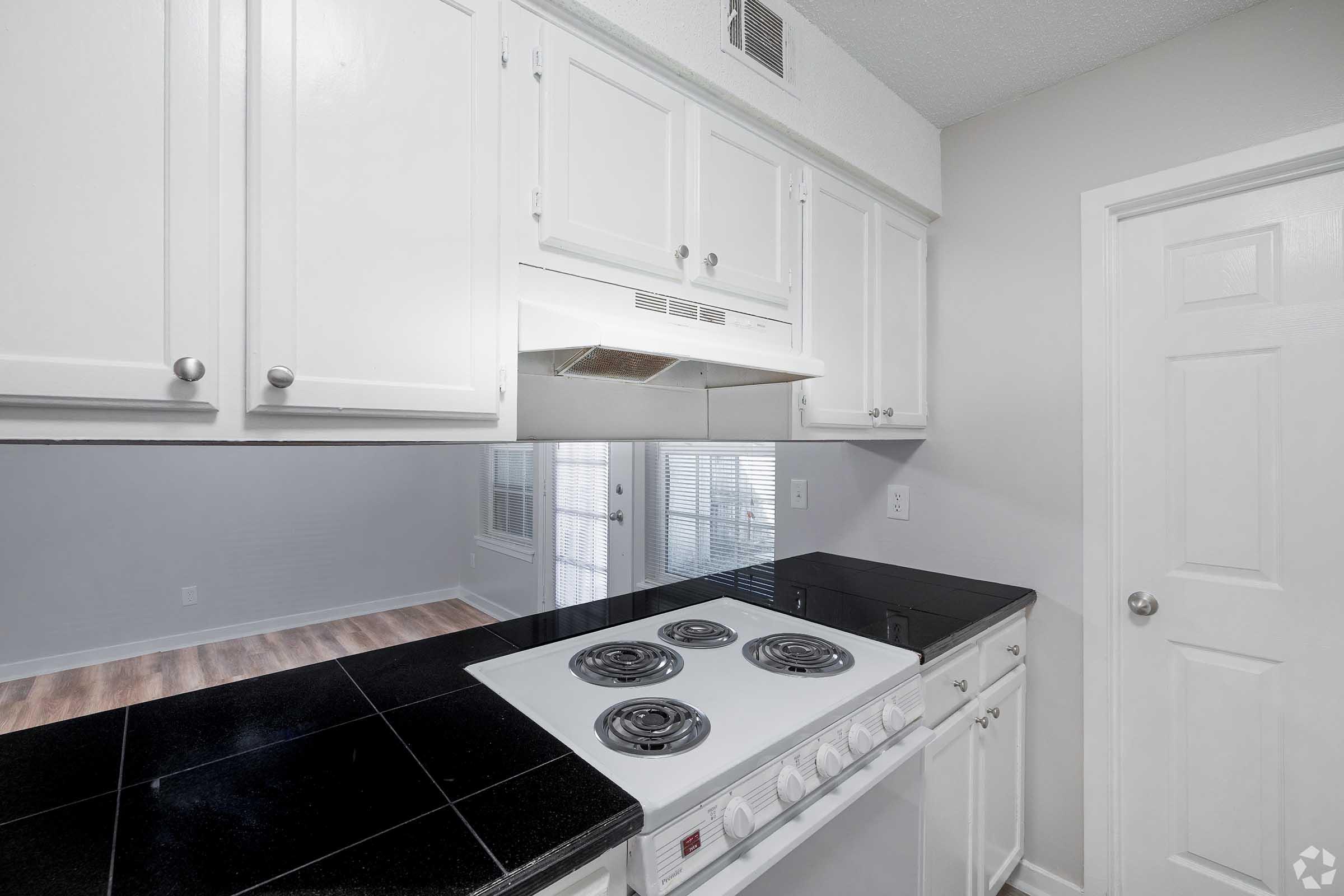 Modern kitchen featuring white cabinetry, a black countertop, and a stovetop with four burners. There's a range hood above the stove, and the wall behind it is partially visible. A door on the right leads to another room, and large windows create a bright atmosphere. The flooring is light-colored wood.