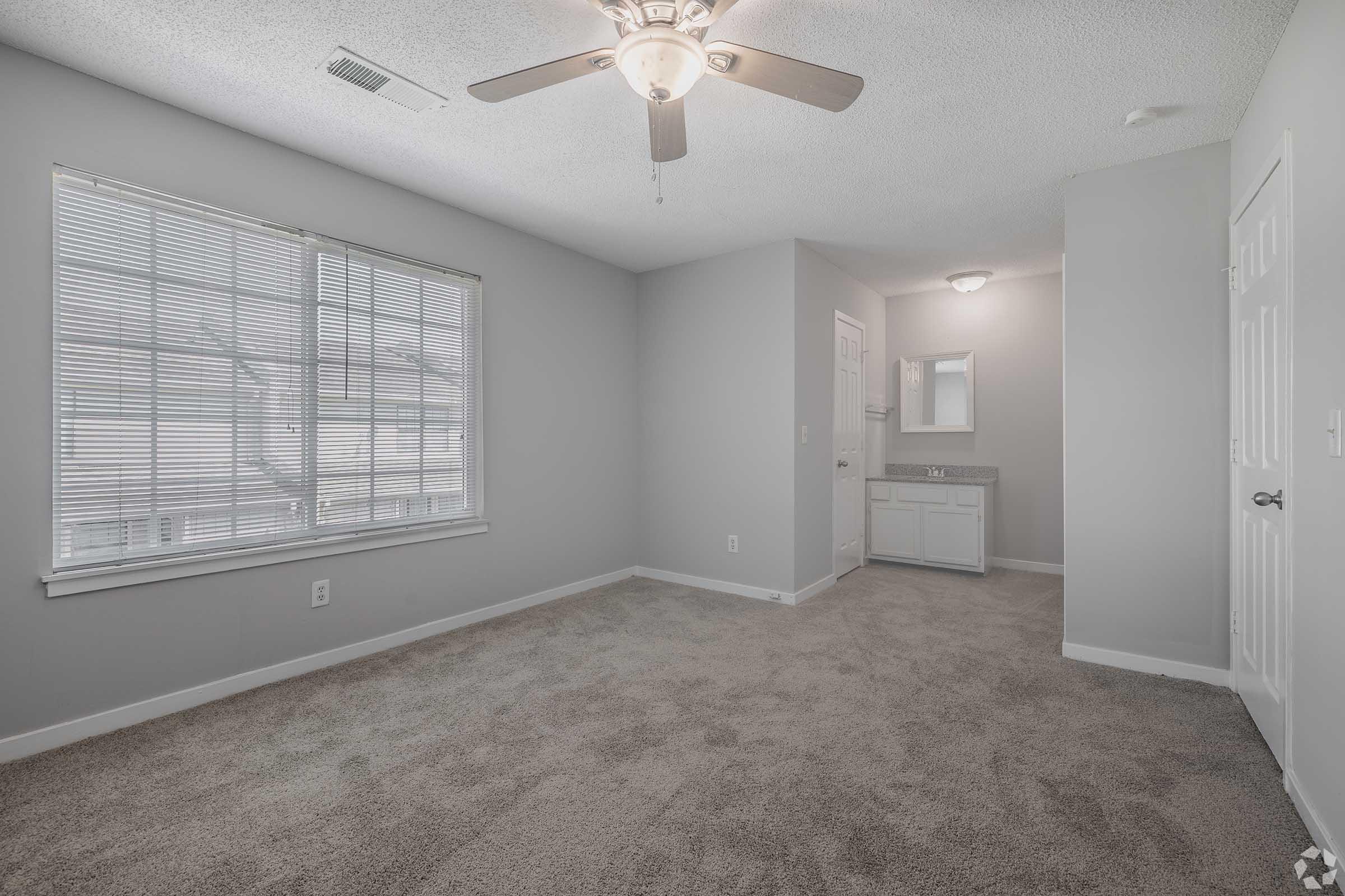 A bright, empty room with light gray walls and beige carpet. It features a ceiling fan, a large window with blinds, and a corner that includes a bathroom vanity area with a mirror. Natural light filters into the space, creating an inviting atmosphere.