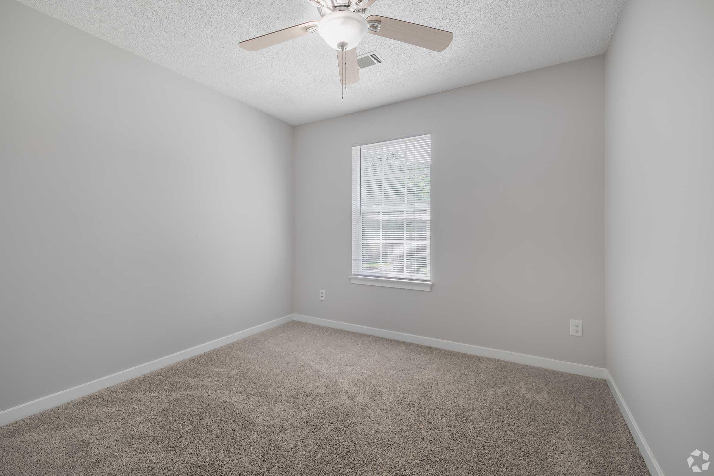 A vacant room with light gray walls and carpeted flooring. There's a ceiling fan with light fixtures and a window allowing natural light to enter. The space appears clean and uncluttered, ideal for personal customization or furniture arrangement.