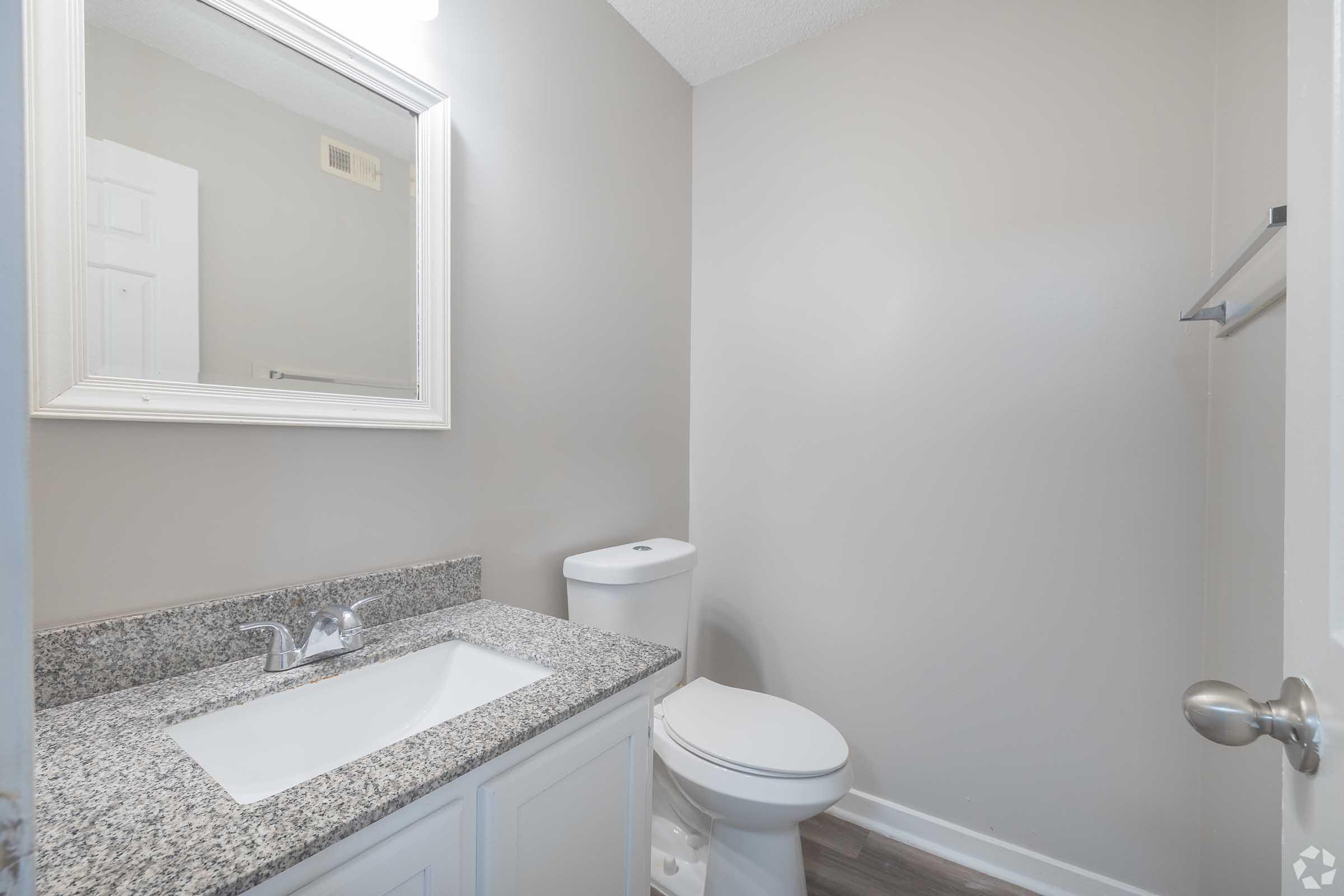 A clean, modern bathroom featuring a granite countertop sink with a mirror above, a white toilet, and a light gray wall. The room is well-lit with natural light coming from an unspecified source, and a towel rack is mounted on the wall. The overall aesthetic is simple and contemporary.