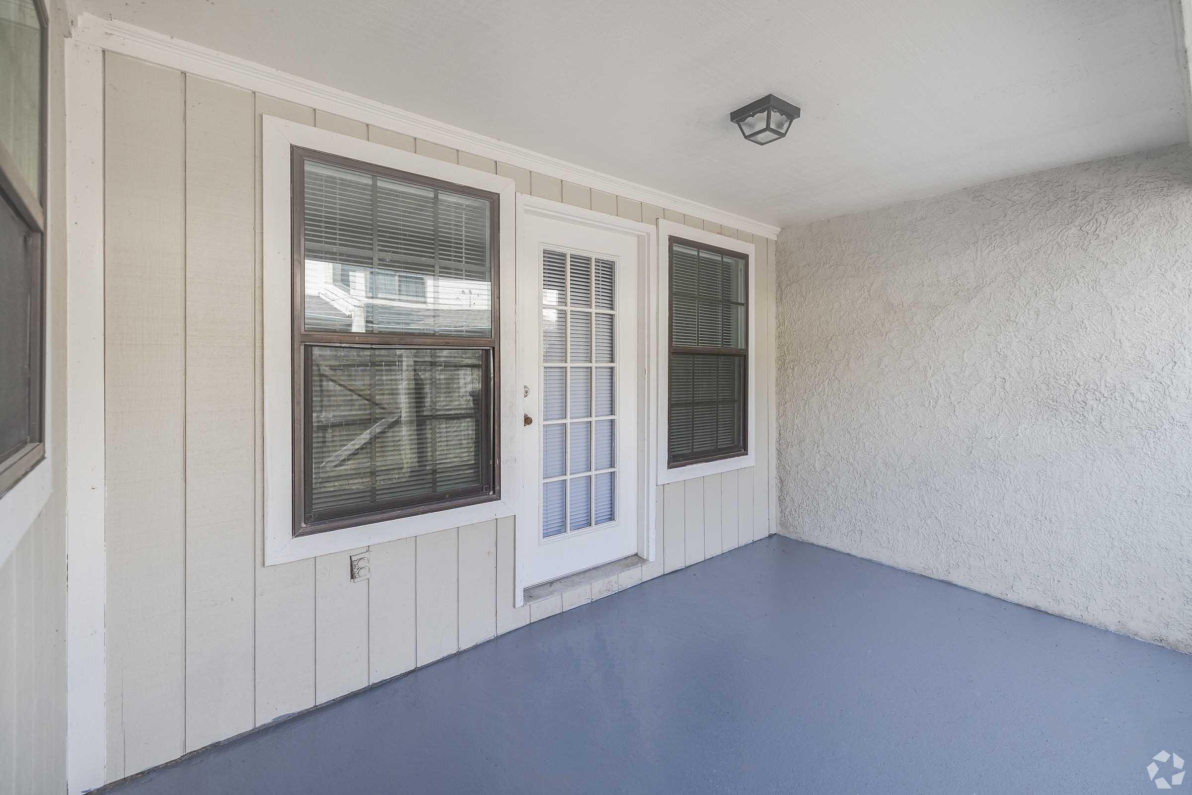 An enclosed porch featuring two large windows and a white door in the center. The floor is painted gray, and the walls are a light beige with textured finish. A ceiling light fixture is visible. The space is well-lit and offers a cozy outdoor view.