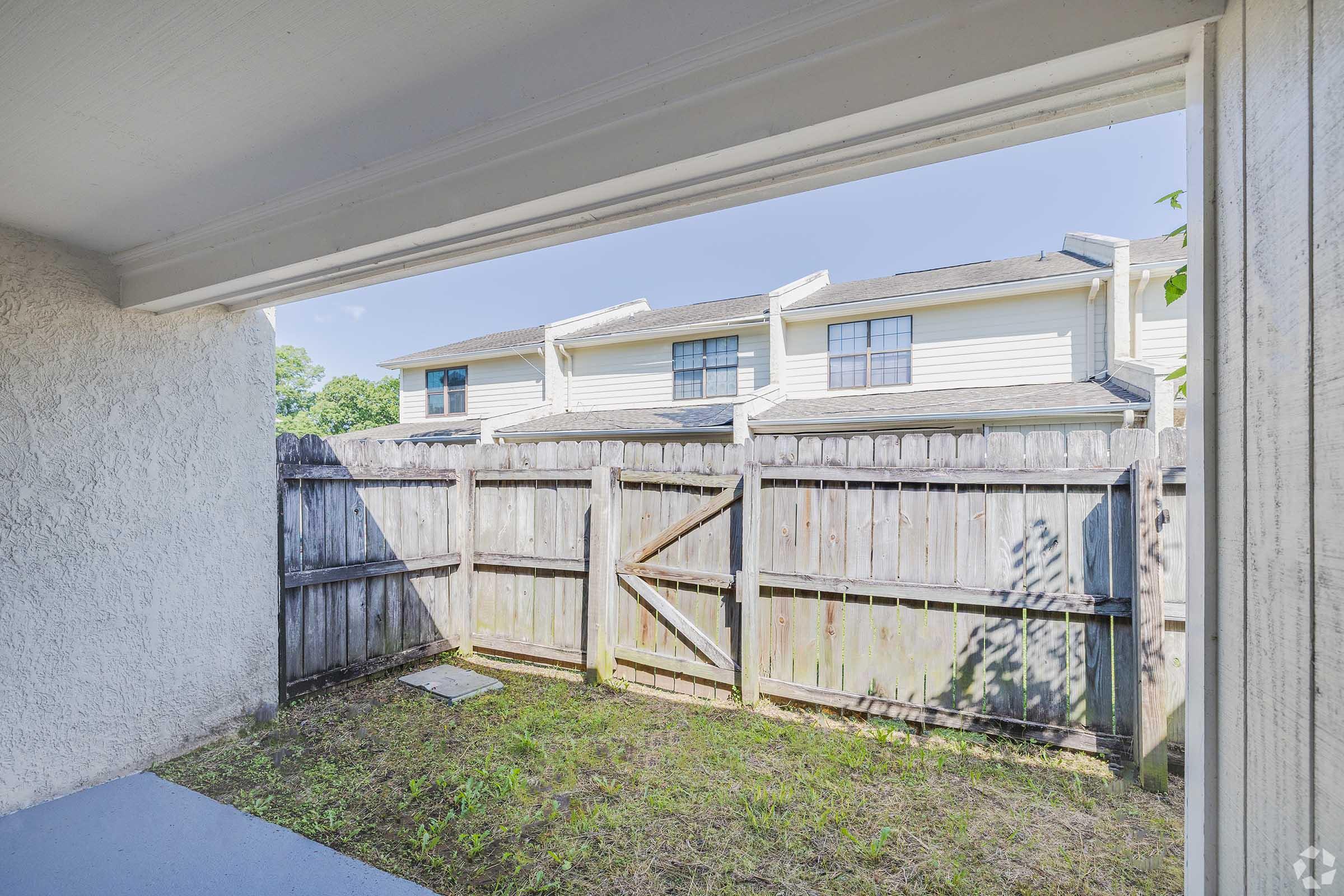 View from a patio or porch showing a fenced yard with grass and a small slab of concrete. In the background, residential buildings can be seen with windows and a roofline. The scene is bright and sunny, suggesting a warm day.