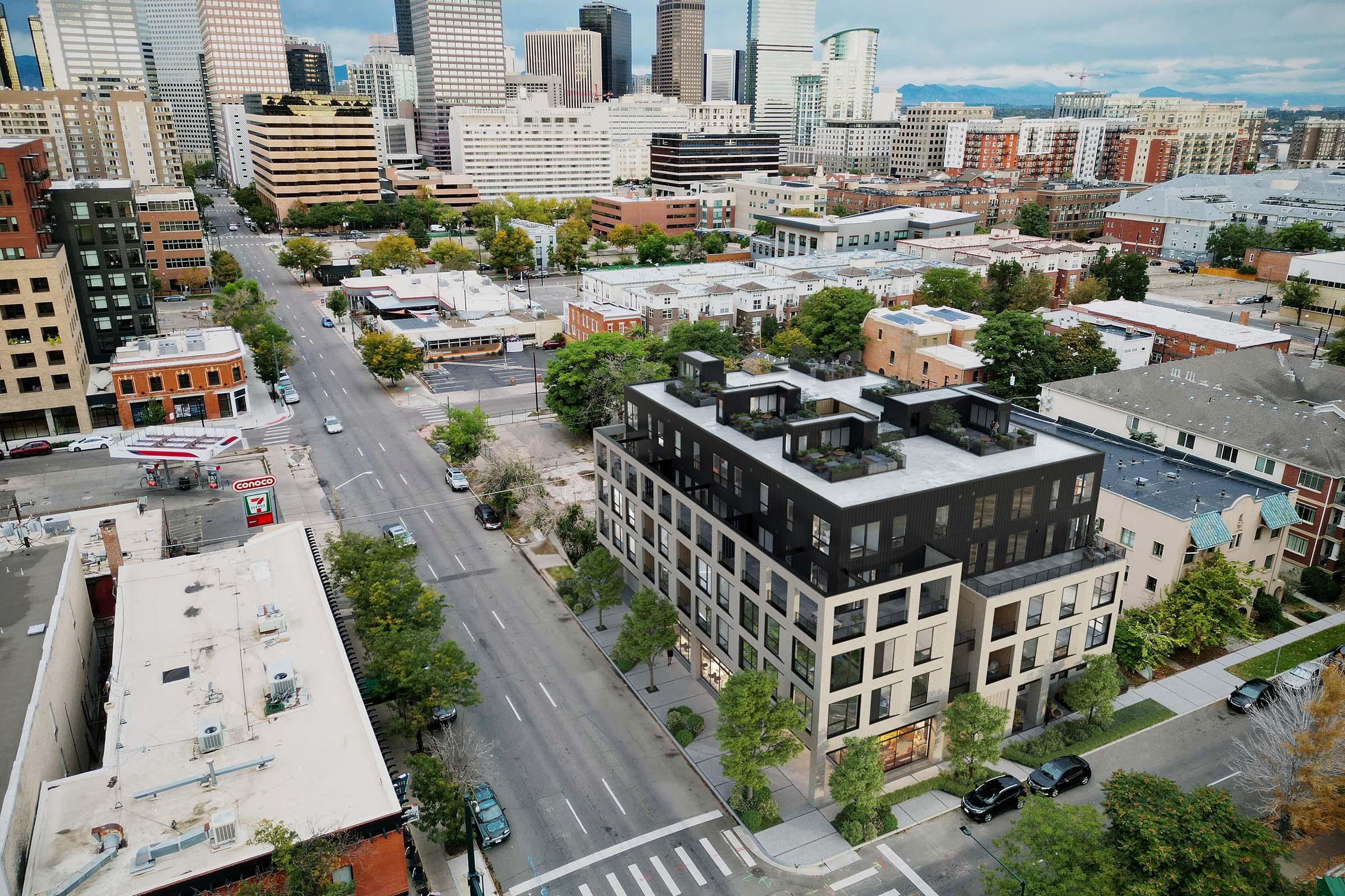 Aerial view of a modern building in an urban area, surrounded by a mix of low-rise and high-rise structures. The scene includes tree-lined streets, parked cars, and city skyscrapers in the background under a cloudy sky.