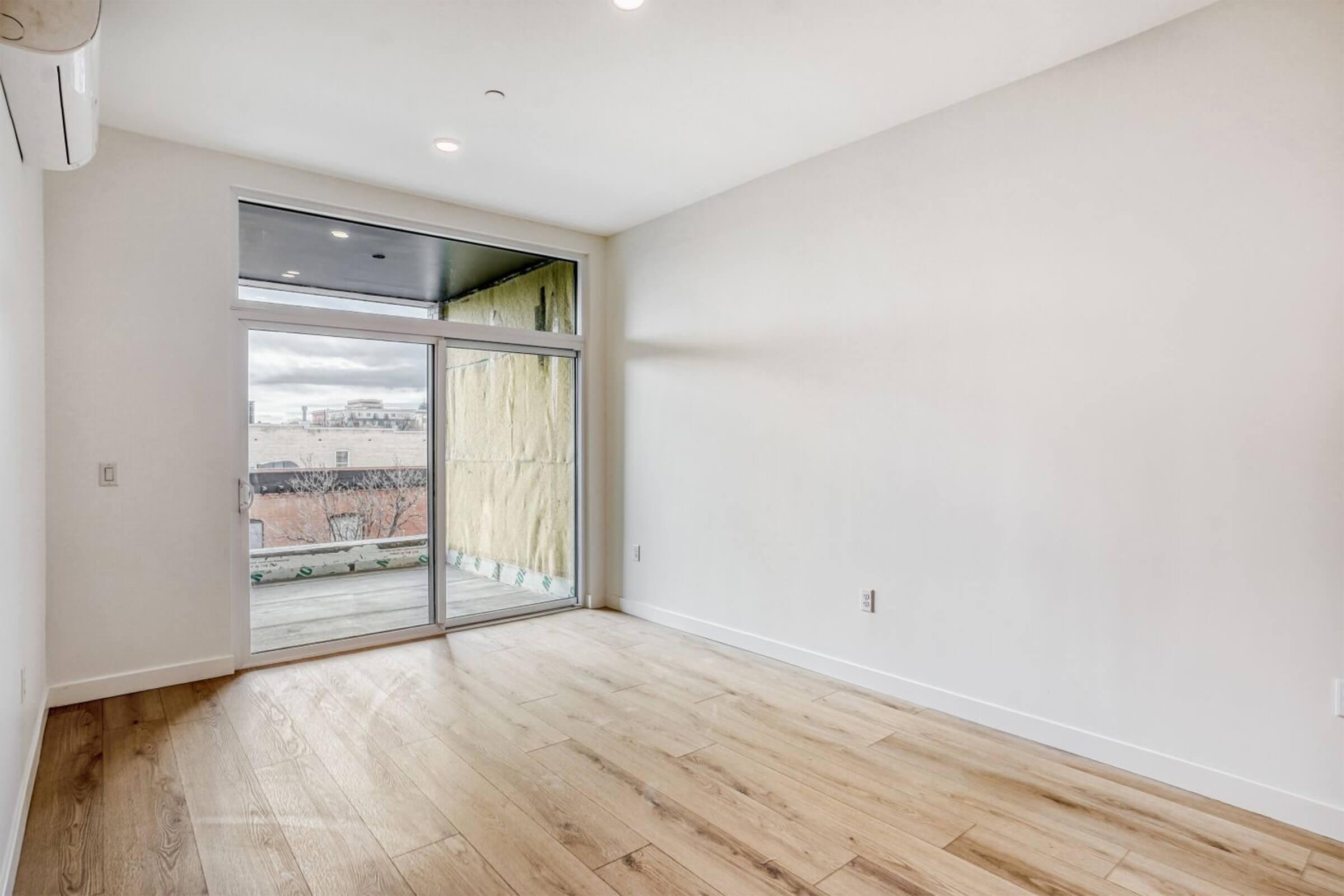 A spacious, empty room with hardwood flooring, featuring a large sliding glass door that opens to a balcony. The walls are light-colored, and there is natural light coming in. Construction materials are visible through the doorway, indicating ongoing work.