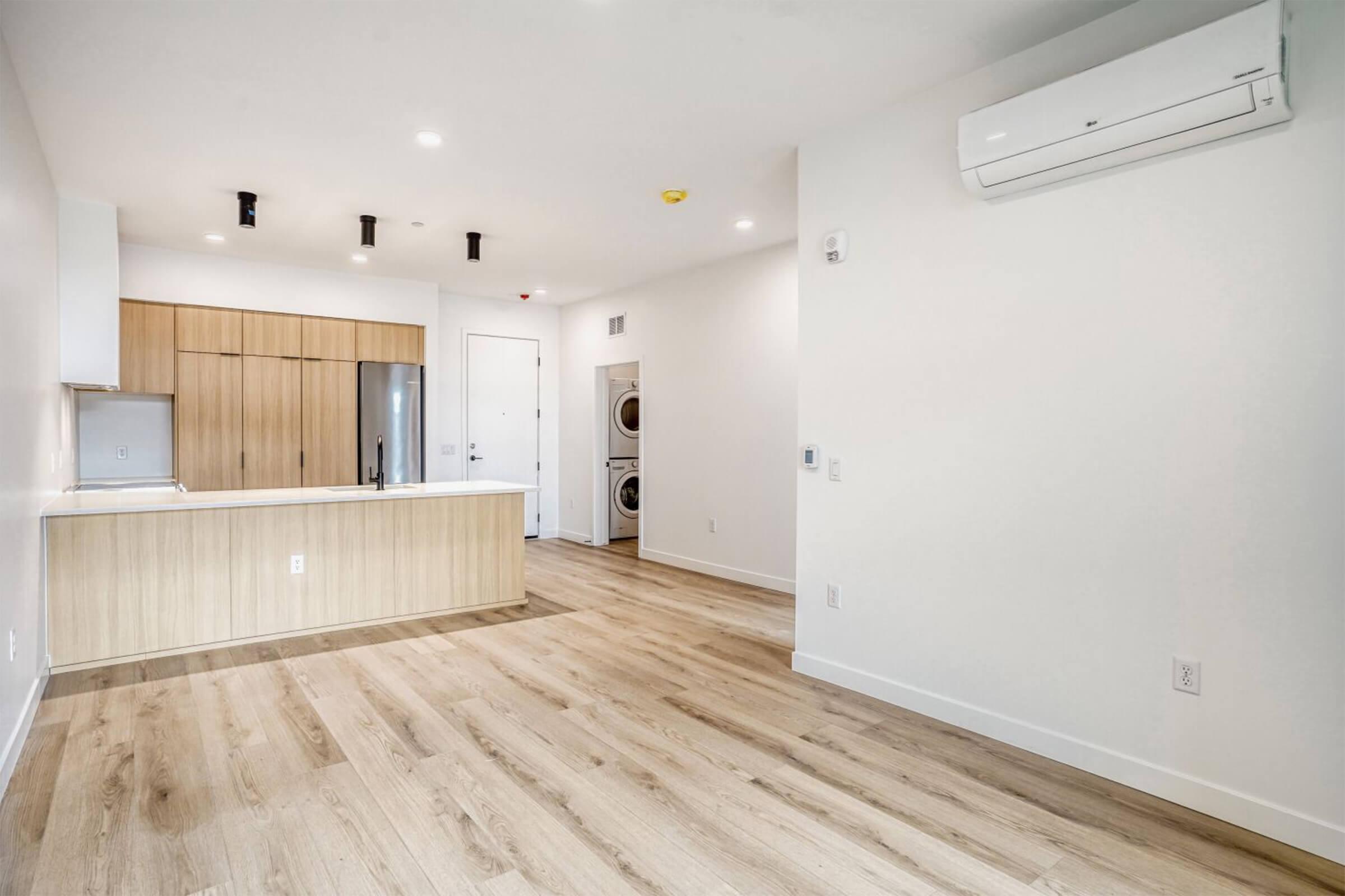 A modern, empty apartment interior featuring a spacious living area with light wood flooring. There is a kitchen area with a light wooden cabinetry and stainless steel appliances. A wall-mounted air conditioning unit is visible, along with a laundry nook with stacked appliances. The walls are painted white, creating a bright atmosphere.