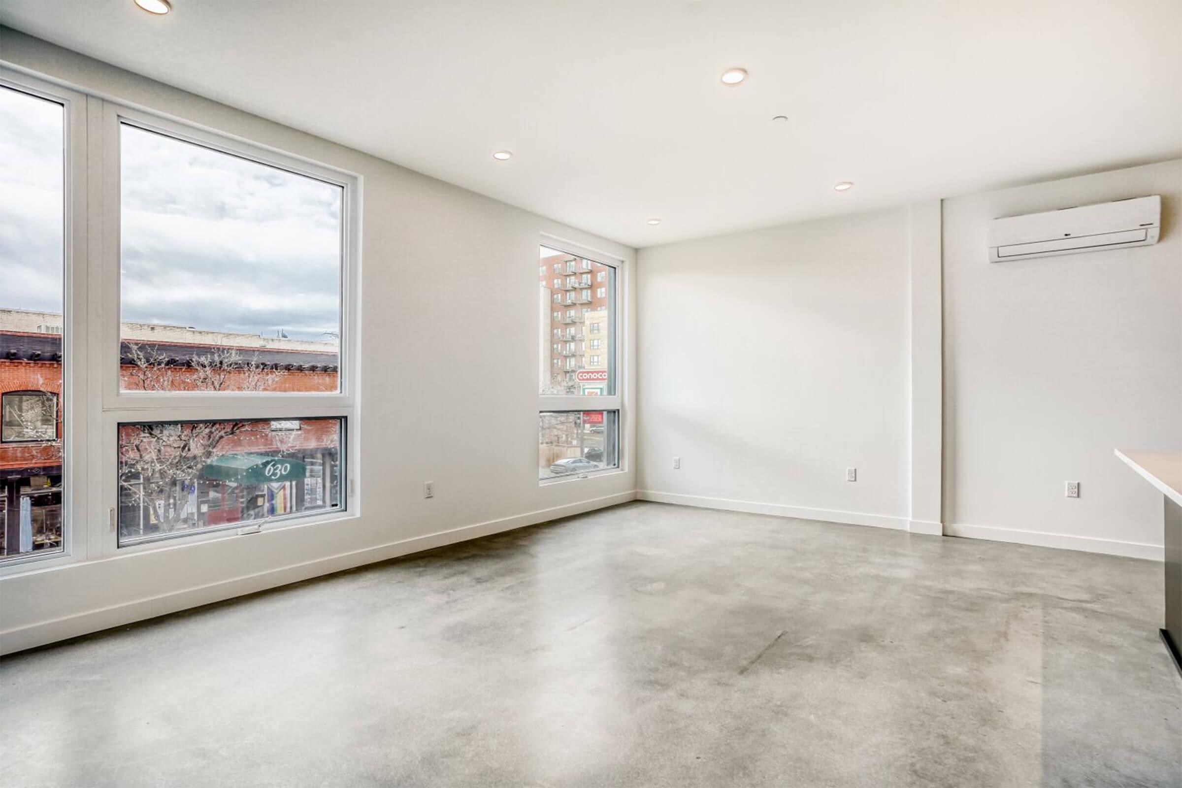 A bright, modern interior of a room featuring polished concrete floors and large windows that provide natural light. The walls are painted white, and there's an air conditioning unit on the wall. The view includes a glimpse of the street outside with buildings in the background.