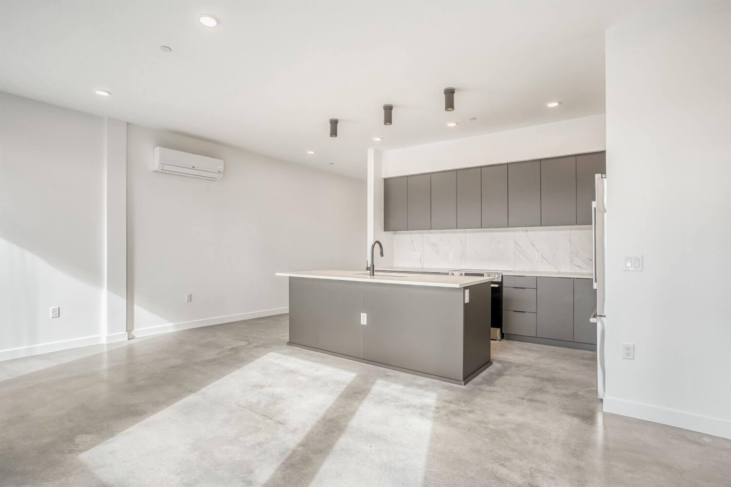 Modern kitchen with a minimalist design, featuring a large island with a sink, dark cabinetry, and light-colored marble accents. The space has concrete flooring and is well-lit with natural light from a nearby window. Air conditioning unit on the wall and sleek appliances complement the contemporary aesthetic.
