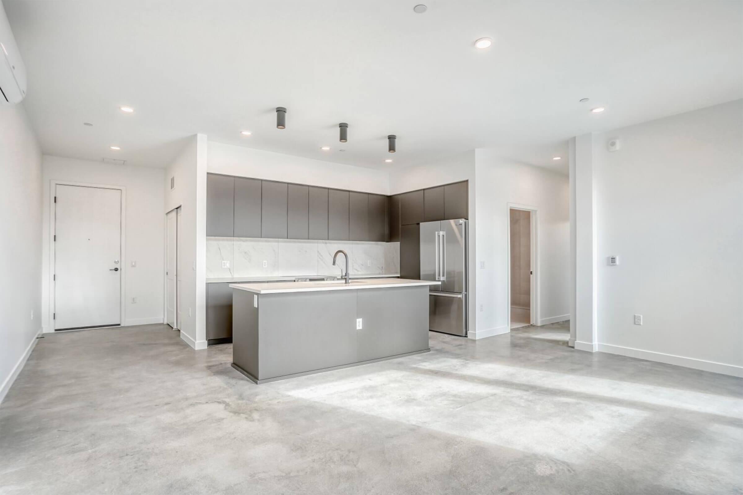 A modern open-concept kitchen and living area featuring a sleek island with a sink, stainless steel appliances, and gray cabinetry. The space has polished concrete flooring, white walls, and several ceiling lights, creating a bright and inviting atmosphere.