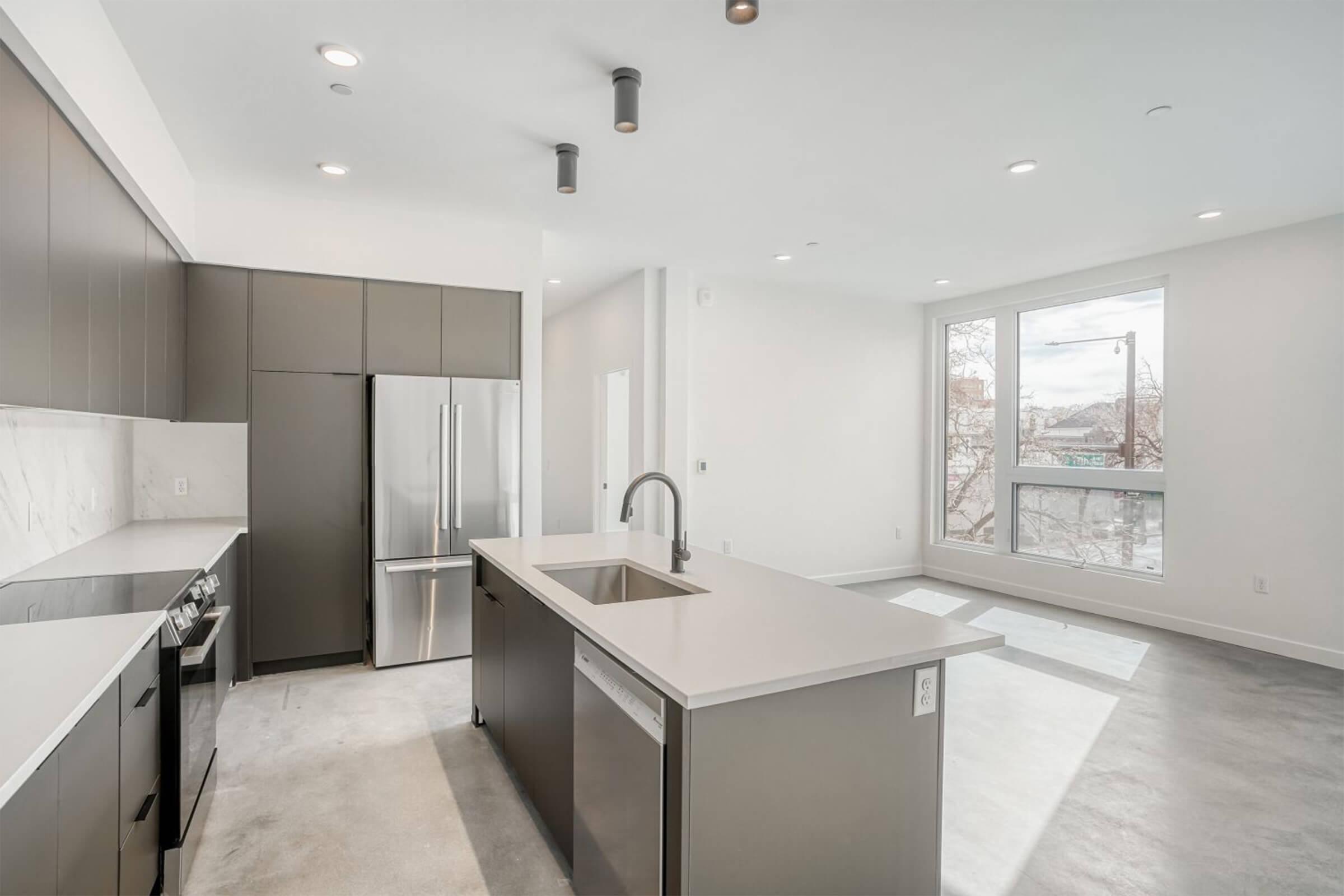 Modern kitchen with sleek dark cabinetry, stainless steel appliances, and a large island with a light countertop. Natural light floods in from a large window, illuminating the open space with polished concrete floors and minimalist design elements.