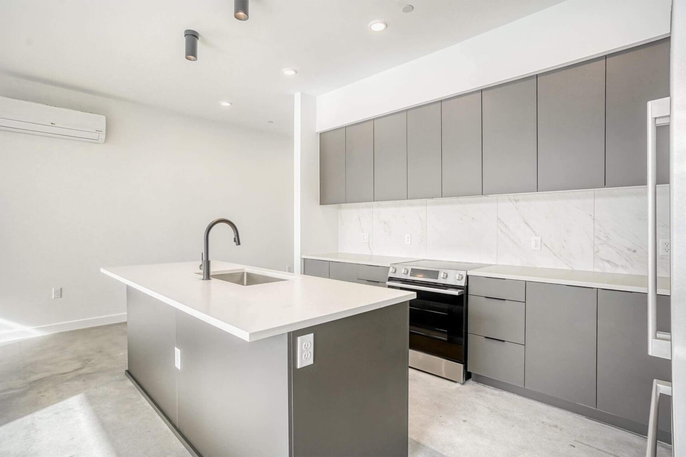 Modern kitchen featuring a central island with a sink, sleek gray cabinetry, a black oven, and white countertops. The space is bright with natural light, minimalistic design, and an air conditioning unit on the wall. The floor is concrete, giving a contemporary aesthetic.