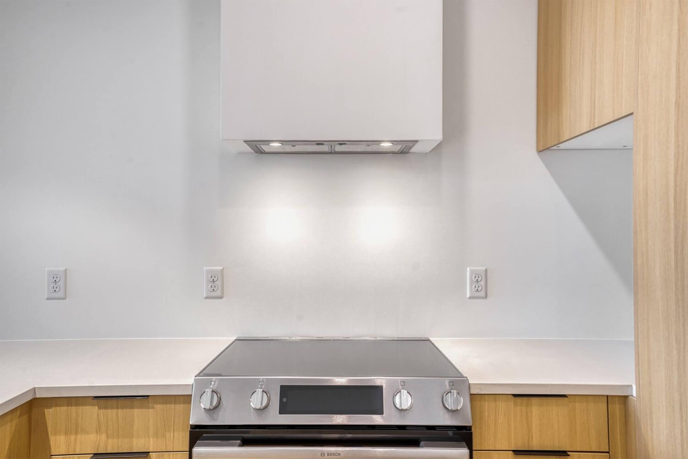 A modern kitchen featuring a sleek stove with a stainless steel oven below it, a minimalist range hood above, and wooden cabinetry on either side. The countertop is light-colored, and the walls are painted white, creating a clean and airy atmosphere.