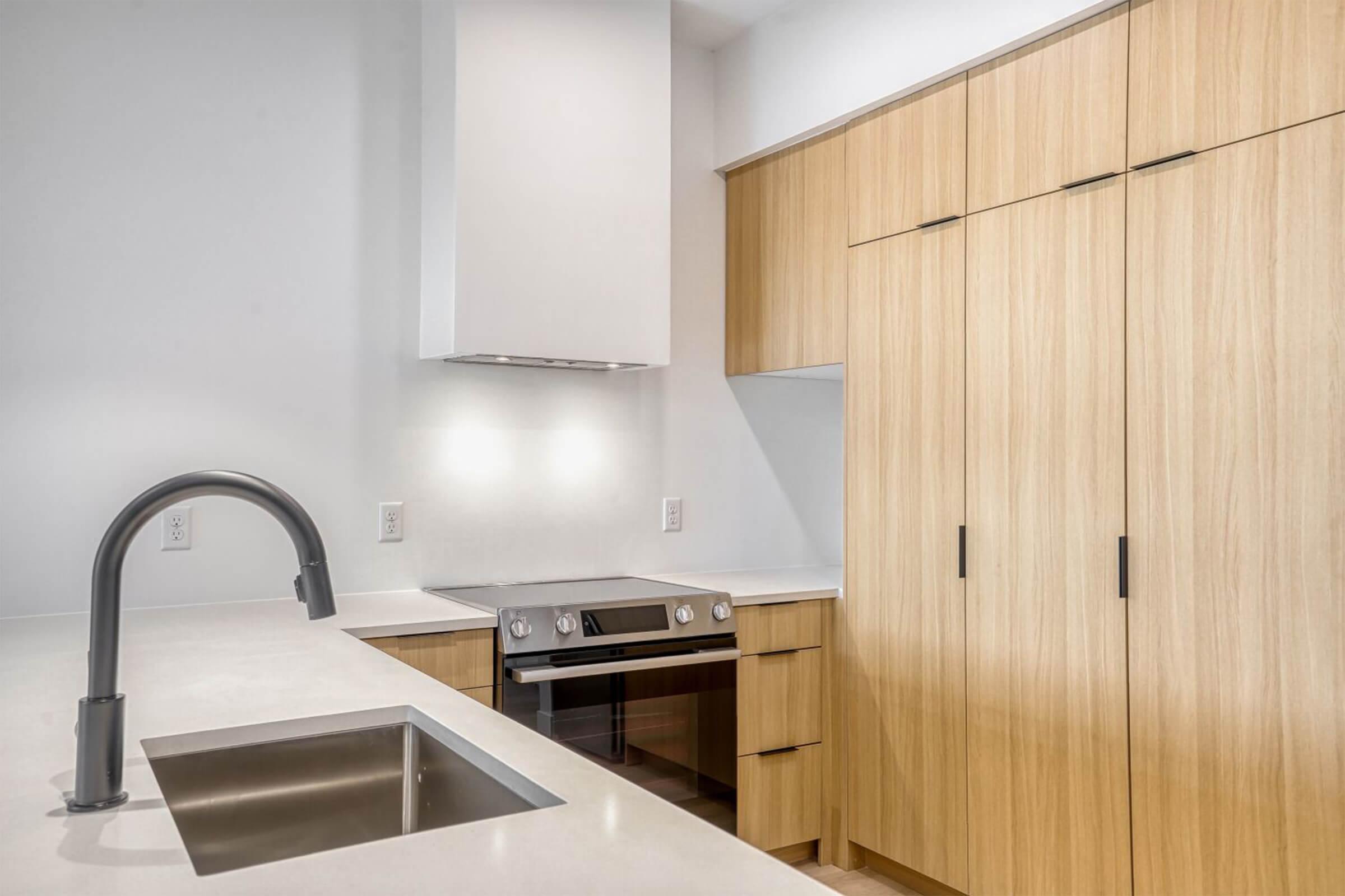 Modern kitchen design featuring a sleek countertop with a built-in sink, a stainless steel oven, and minimalist cabinetry in light wood. The space has white walls and a clean, contemporary aesthetic, illuminated by soft lighting. Ideal for cooking and entertaining.