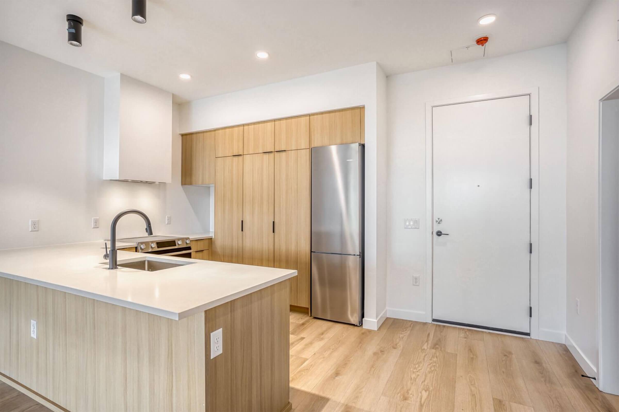 A modern kitchen featuring light wood cabinetry, a central island with a sink, sleek stainless steel appliances, and bright overhead lighting. The space has a clean, minimalist design with neutral colors and smooth finishes, and a doorway leading to an entryway.