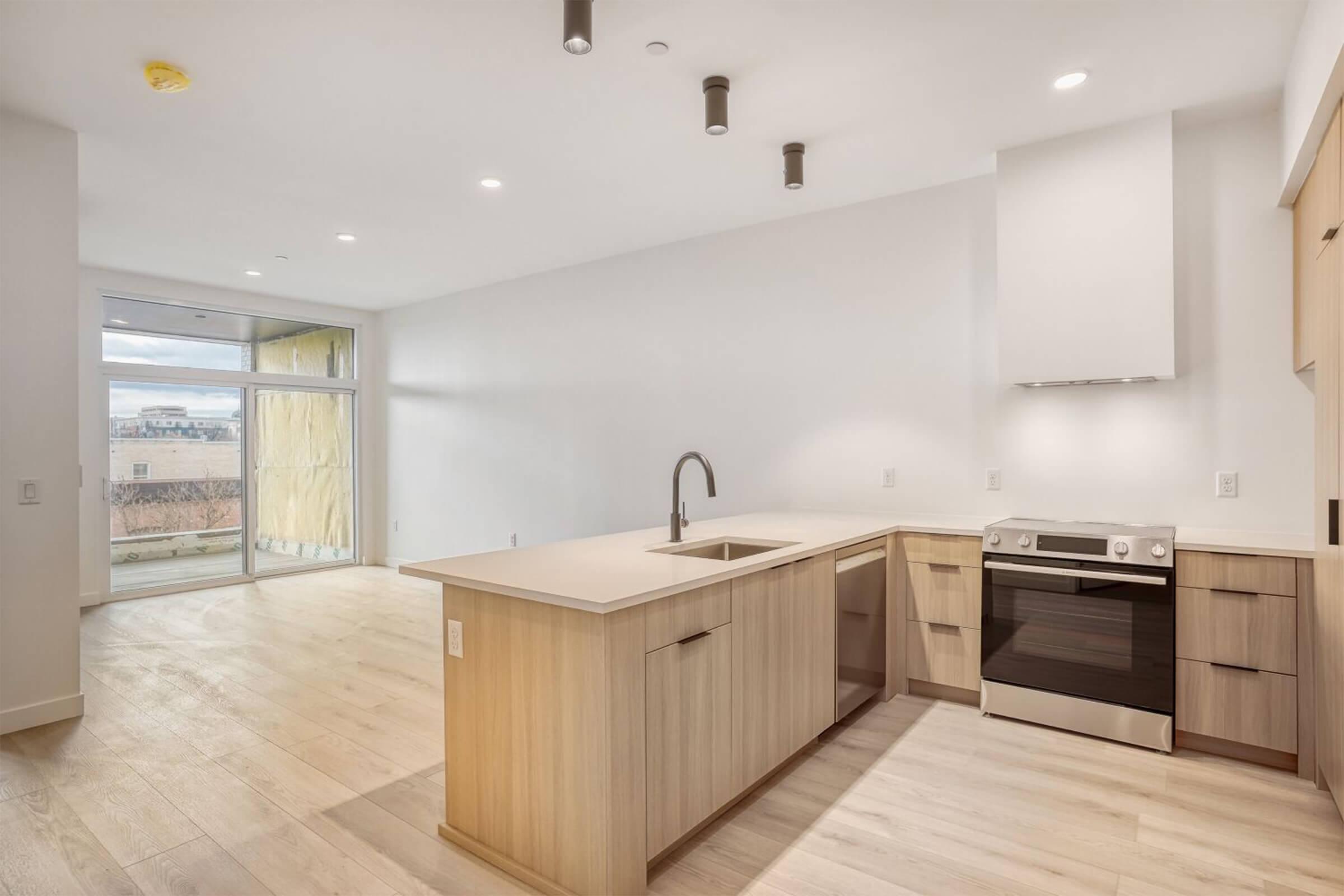 Modern kitchen with light wood cabinetry, a central island with a sink, and a stainless steel stove. The space features an open layout and large windows, providing natural light. Neutral-colored walls and light wood flooring enhance the contemporary feel of the room. View of an outdoor area visible through the windows.