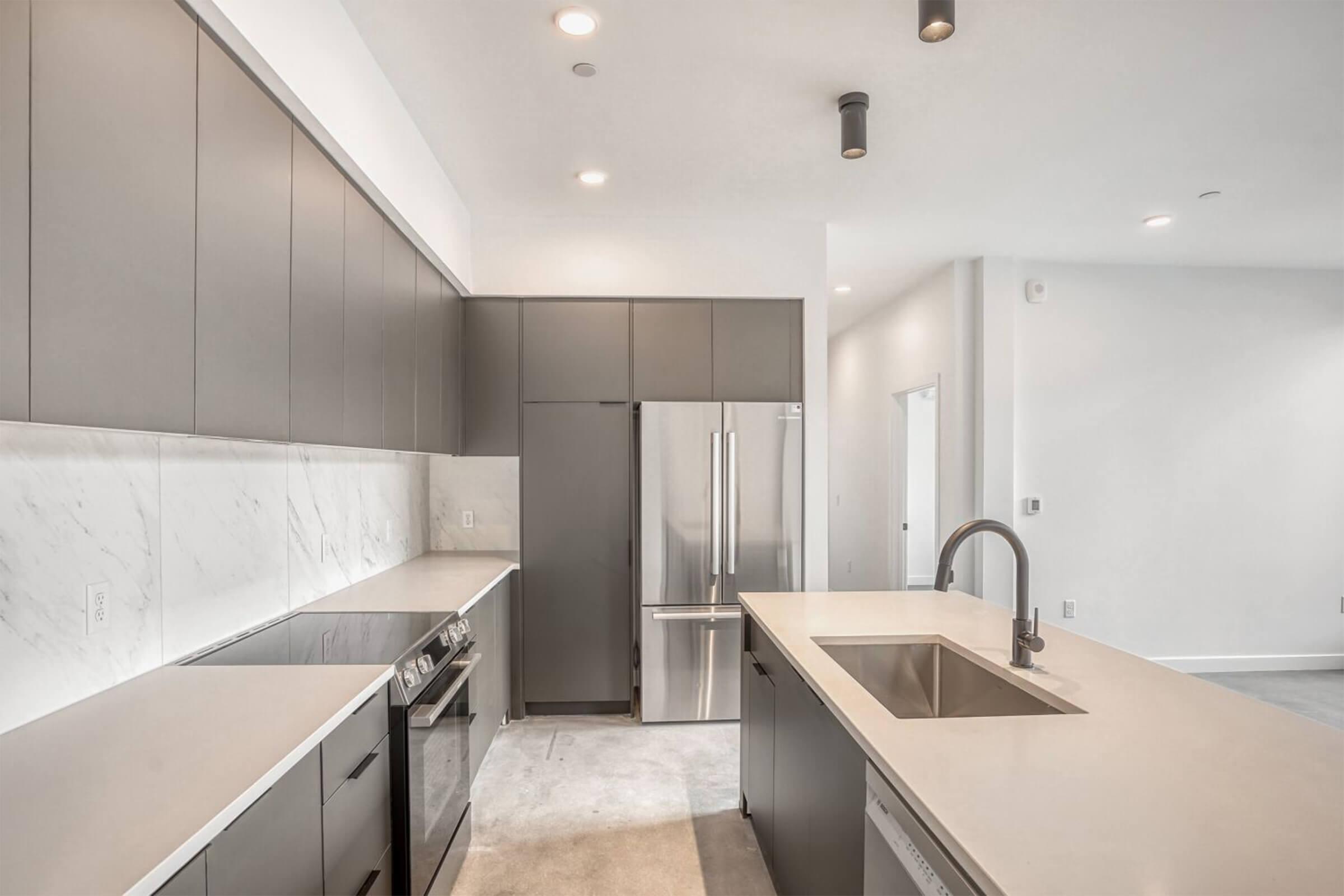 Modern kitchen featuring sleek grey cabinetry, stainless steel appliances, and a large island with a built-in sink. The countertops are light-colored with a minimalist design, and the space is well-lit with recessed lighting. The overall aesthetic is contemporary and clean.