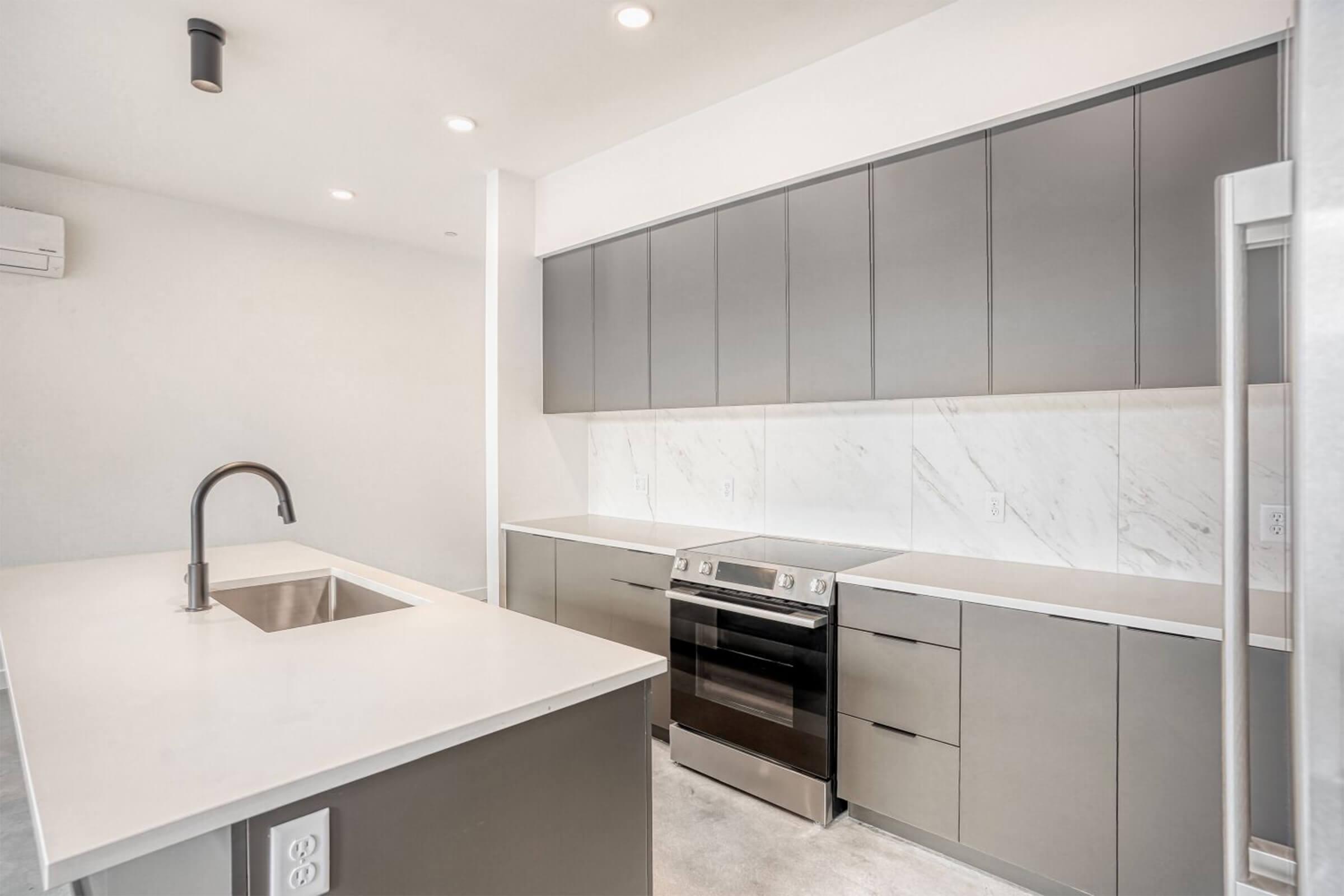 Modern kitchen featuring sleek gray cabinets, a white countertop, and stainless steel appliances. A large island with a built-in sink is in the foreground, while the oven is integrated into the cabinetry. The space has bright lighting and a contemporary design, with a minimalist aesthetic.