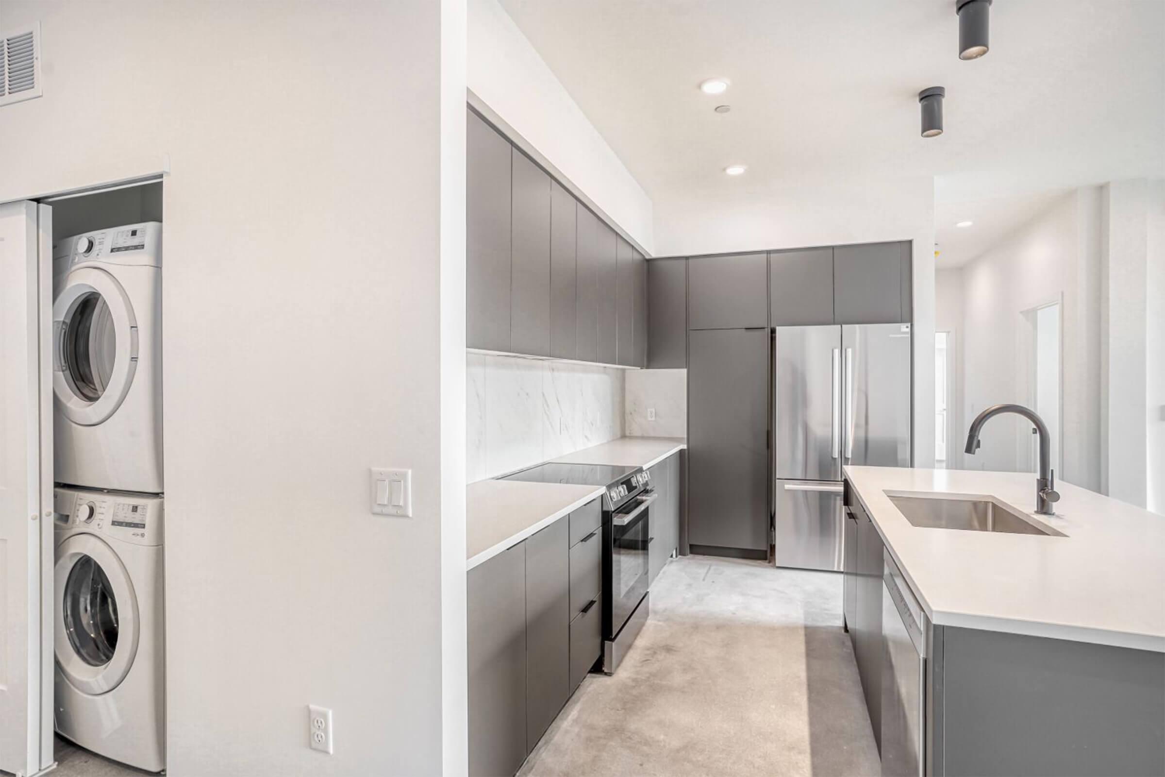 Modern kitchen featuring sleek, dark cabinetry, stainless steel appliances, and a spacious island with a sink. To the left, there is a laundry area with stacked washer and dryer. The space has bright, neutral walls and ample lighting, creating a clean and contemporary atmosphere.