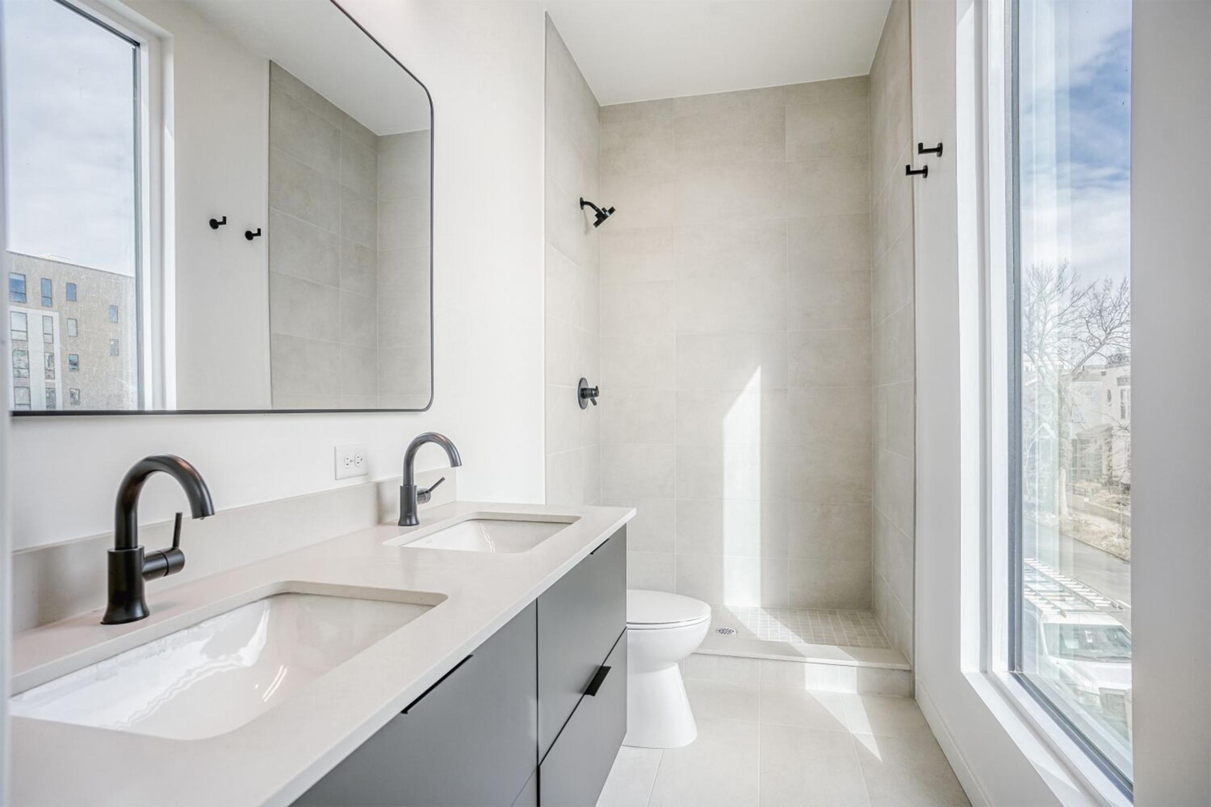 A modern bathroom featuring a double sink with dark faucets, a large mirror above the sinks, and a shower area with a glass wall. The walls are light-colored tiles, and there is a window allowing natural light to enter the space. A sleek toilet is positioned next to the shower.