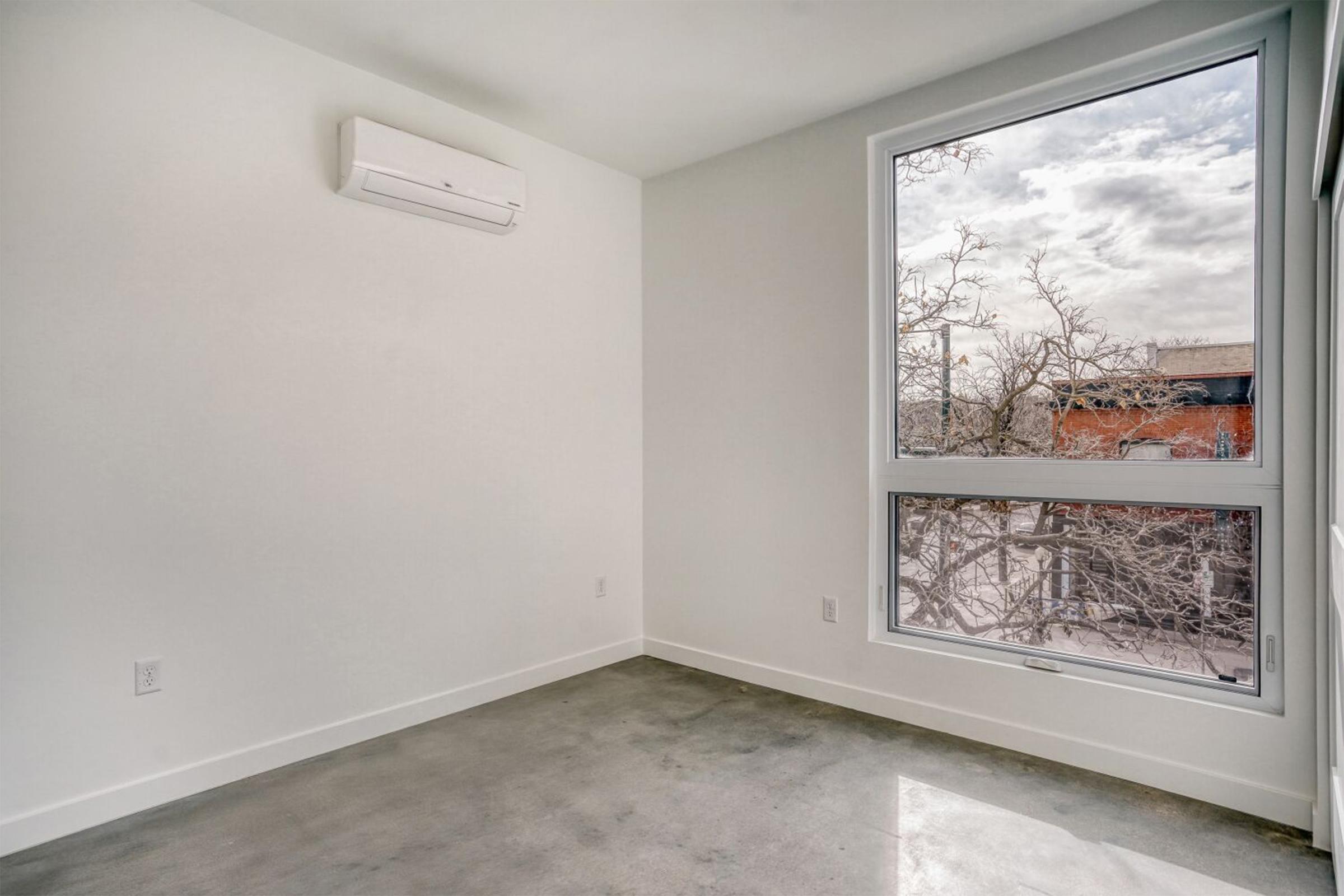 Empty room with light-colored walls and a large window showing a snowy view outside. The floor is made of concrete, and there is a wall-mounted air conditioning unit. Natural light floods the space, highlighting the minimalistic design.