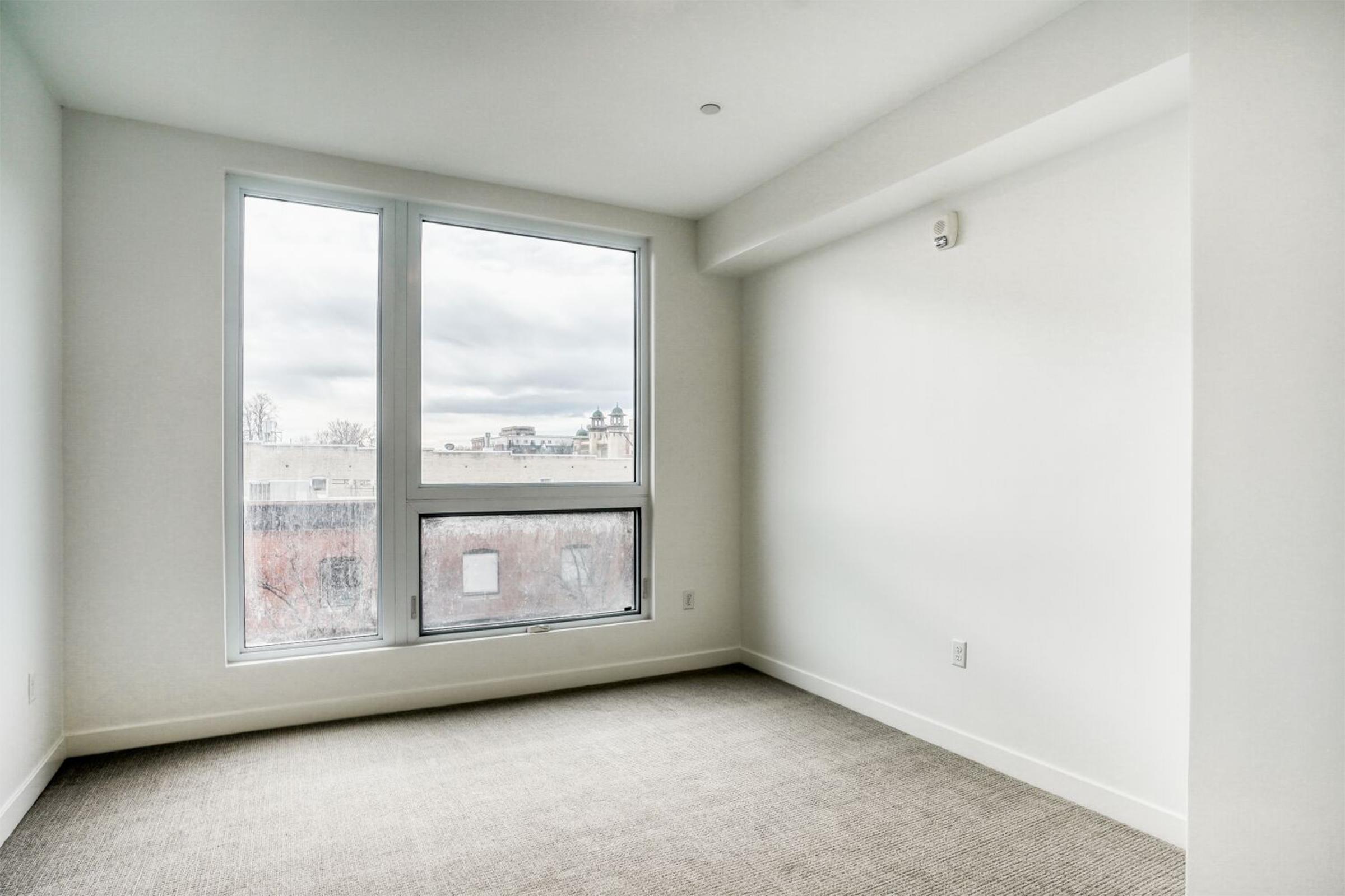 Empty room with large windows letting in natural light, featuring neutral-colored walls and a carpeted floor. The view outside shows a cloudy sky. The space is minimalistic with no furniture or decorations, emphasizing the openness and simplicity of the design.